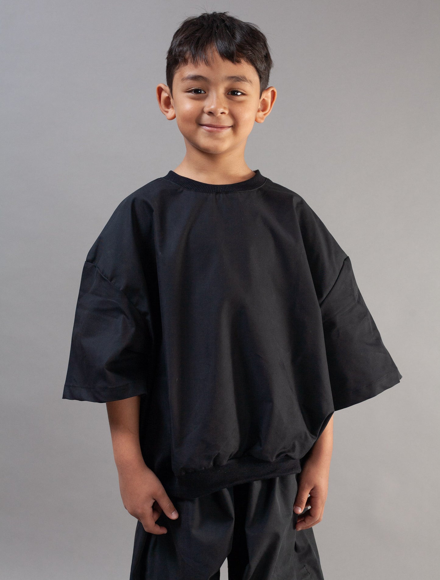 A young boy wearing an oversized black short-sleeve tunic top and matching black pants, standing against a solid gray studio background.
