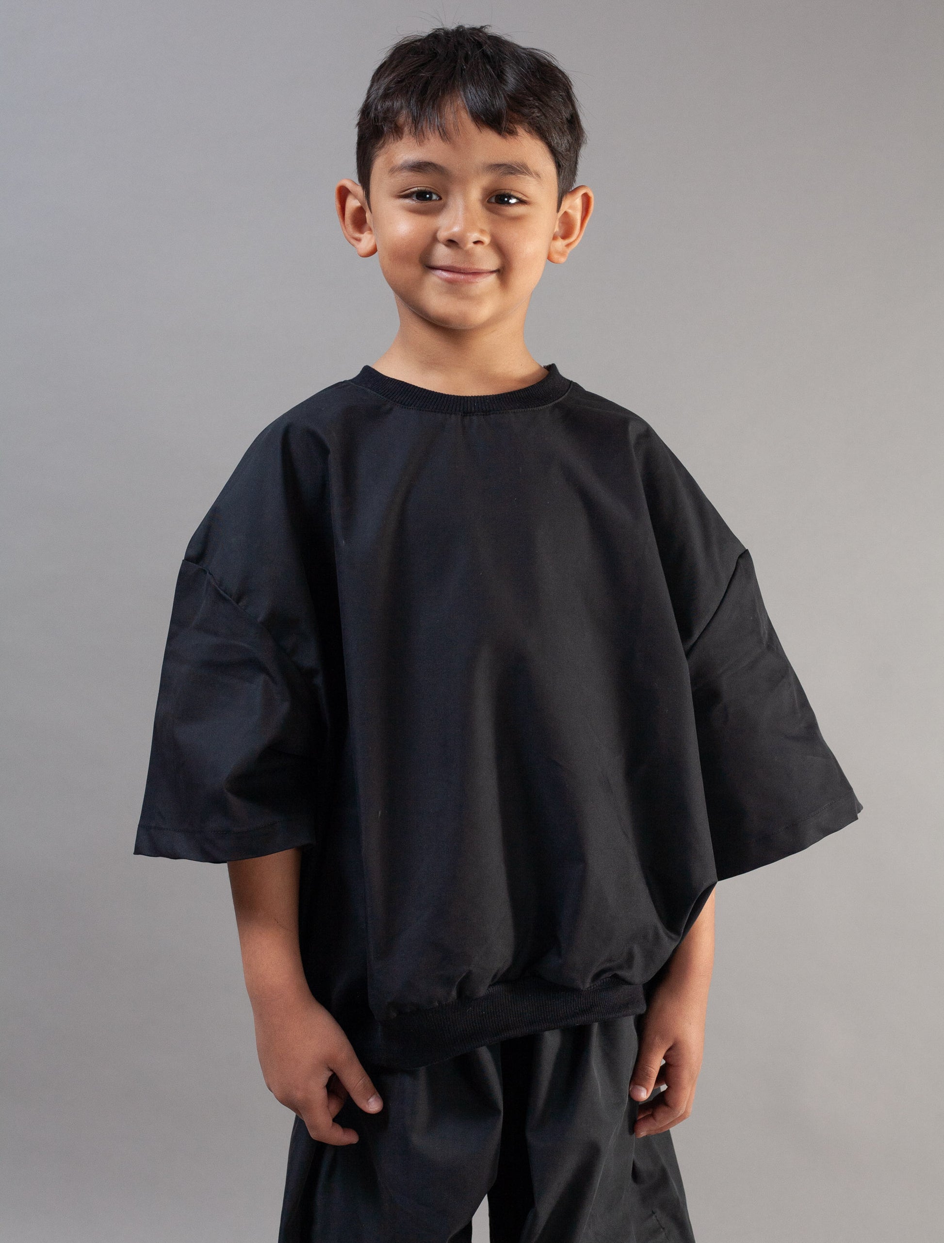 A young boy wearing an oversized black short-sleeve tunic top and matching black pants, standing against a solid gray studio background.