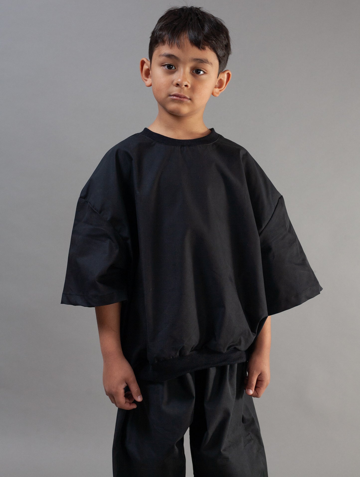 A young boy wearing an oversized black short-sleeve top and matching black pants, standing against a plain gray studio background.