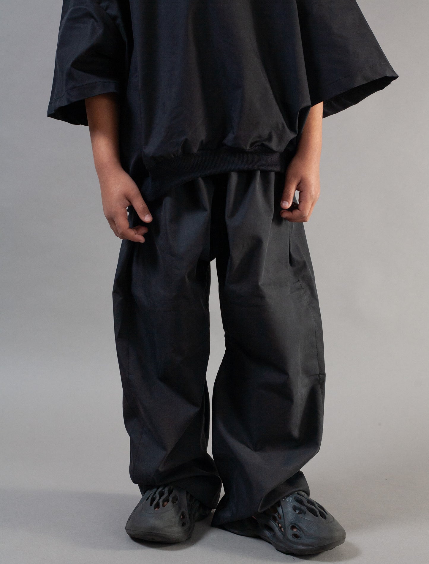 A person wearing loose black wide-leg pants and a matching oversized black top, standing against a plain light gray studio background.