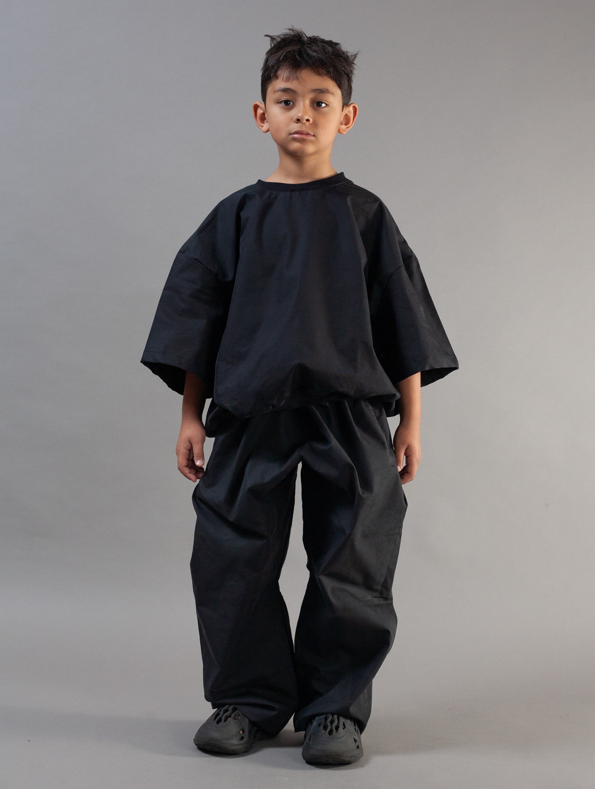 A young boy modeling a black two-piece outfit consisting of a loose-fitting short-sleeve top and matching wide-leg pants, standing against a solid gray studio background.
