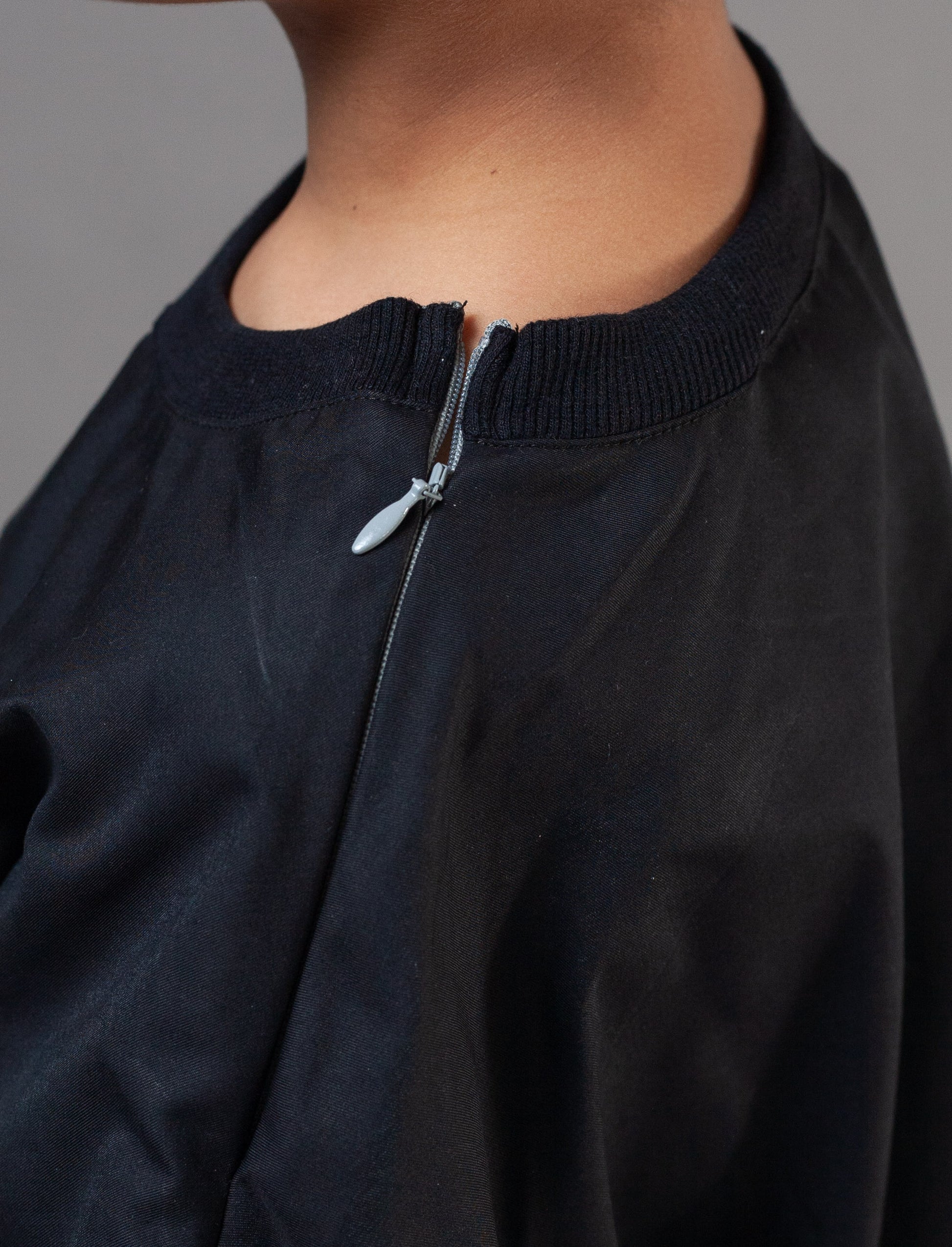 Close-up of a person wearing a black zip-up top with a ribbed crew neck and a small silver zipper pull, against a plain gray background.