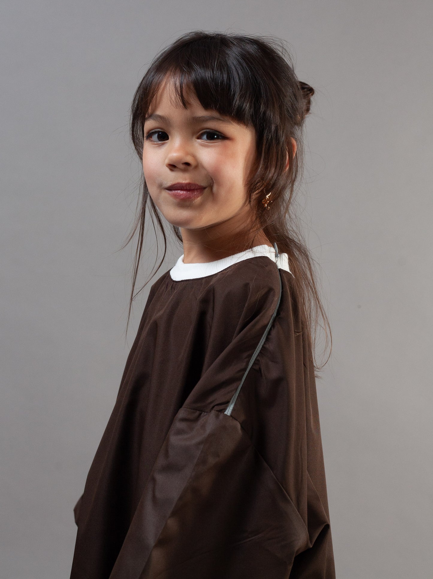 A young girl with dark hair wearing a brown salon cape over a white t-shirt, photographed against a solid gray studio background.