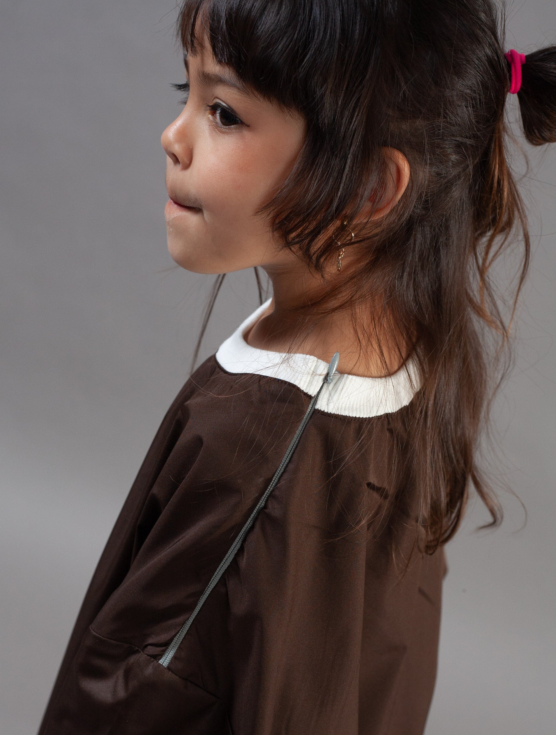 A young girl with dark hair tied back with a pink hair tie is shown in profile wearing a brown jacket with a white collar over a dark top, photographed against a neutral gray background.