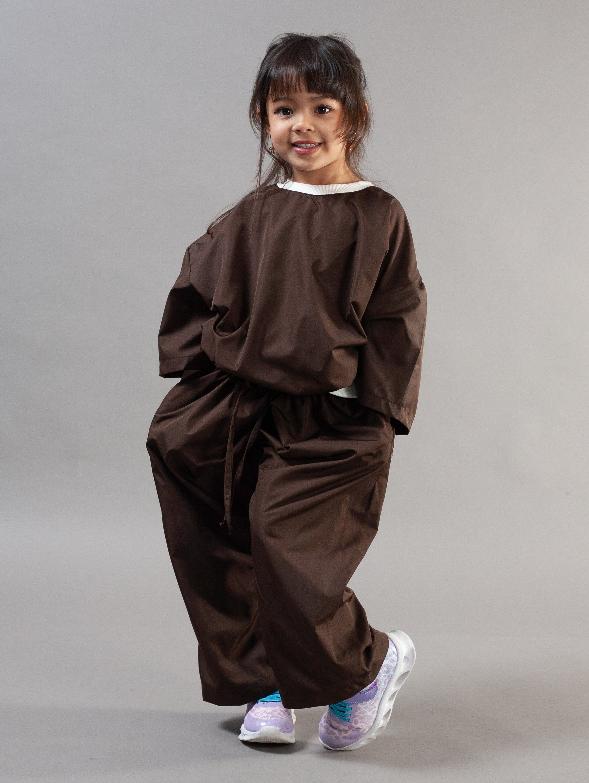 A young girl modeling a dark brown two-piece outfit consisting of a loose long-sleeve top and matching wide-leg pants, paired with white and purple sneakers, against a solid gray studio background.