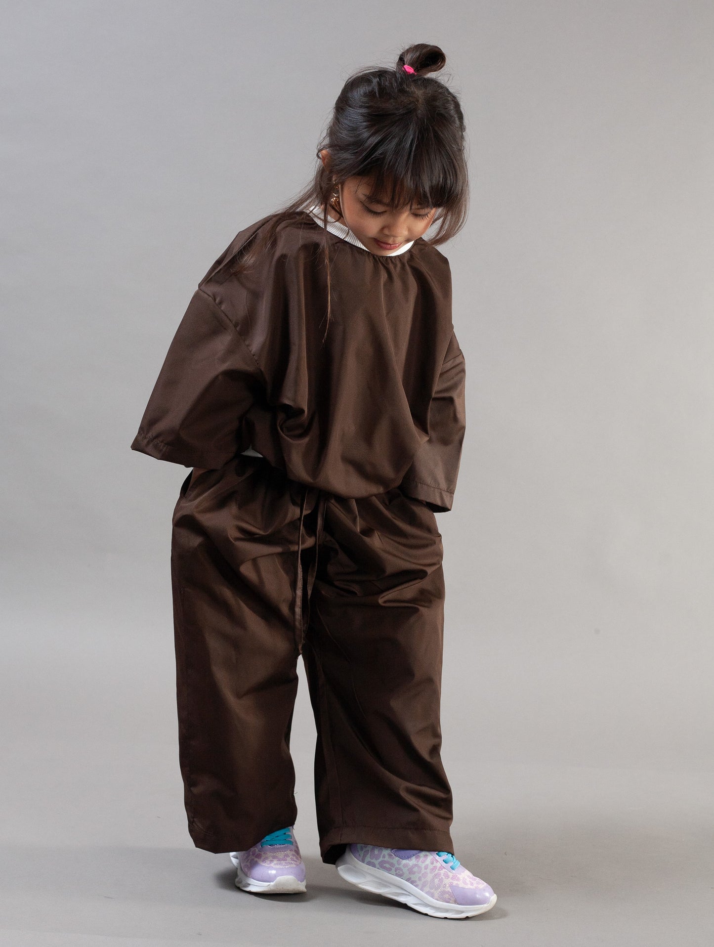 A child wearing an oversized brown hooded sweatshirt and matching loose brown pants, standing against a plain light gray studio background.