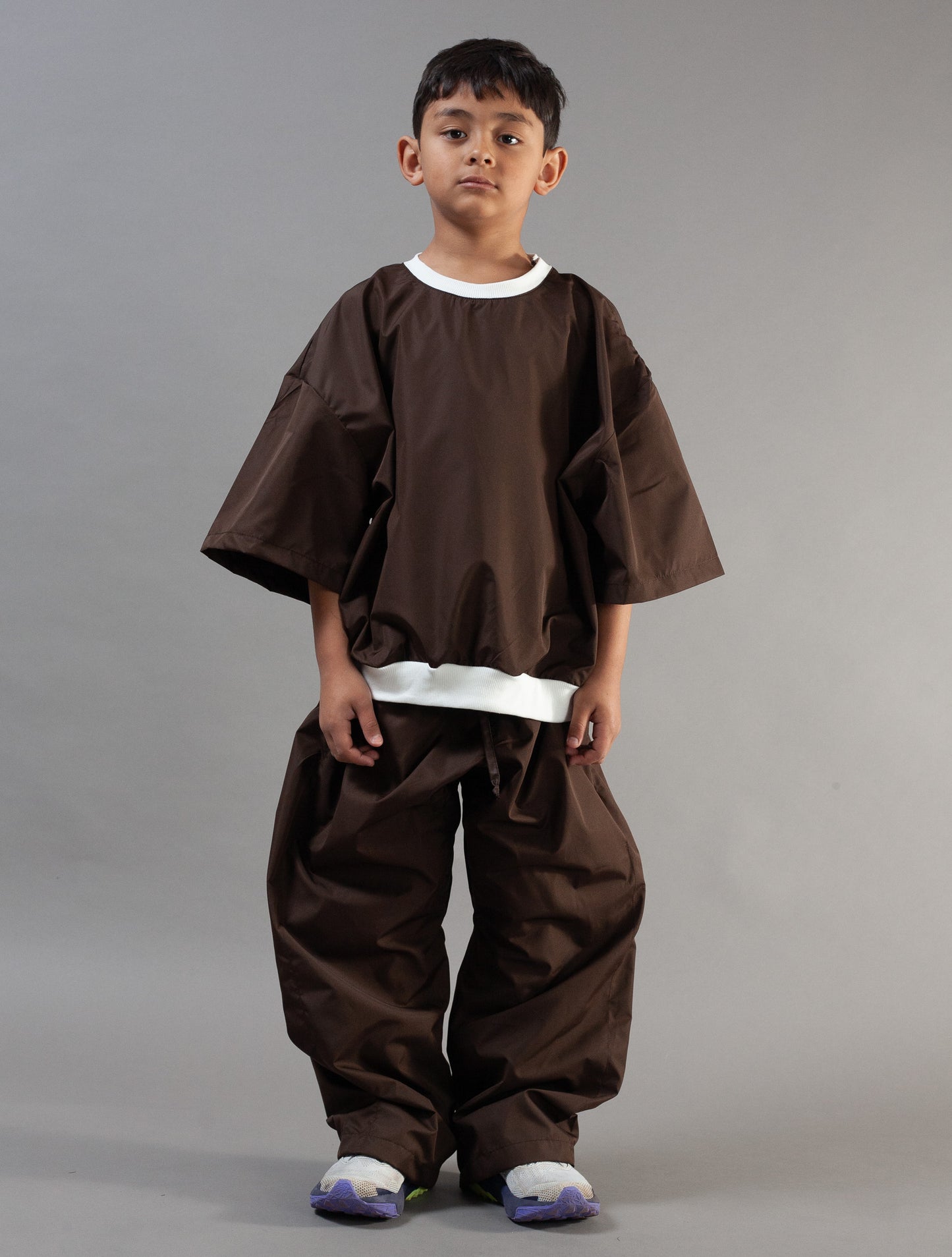 A young boy modeling a dark brown traditional Japanese-style outfit with wide sleeves and loose pants, featuring white contrast trim at the collar and hem, against a solid gray studio background.