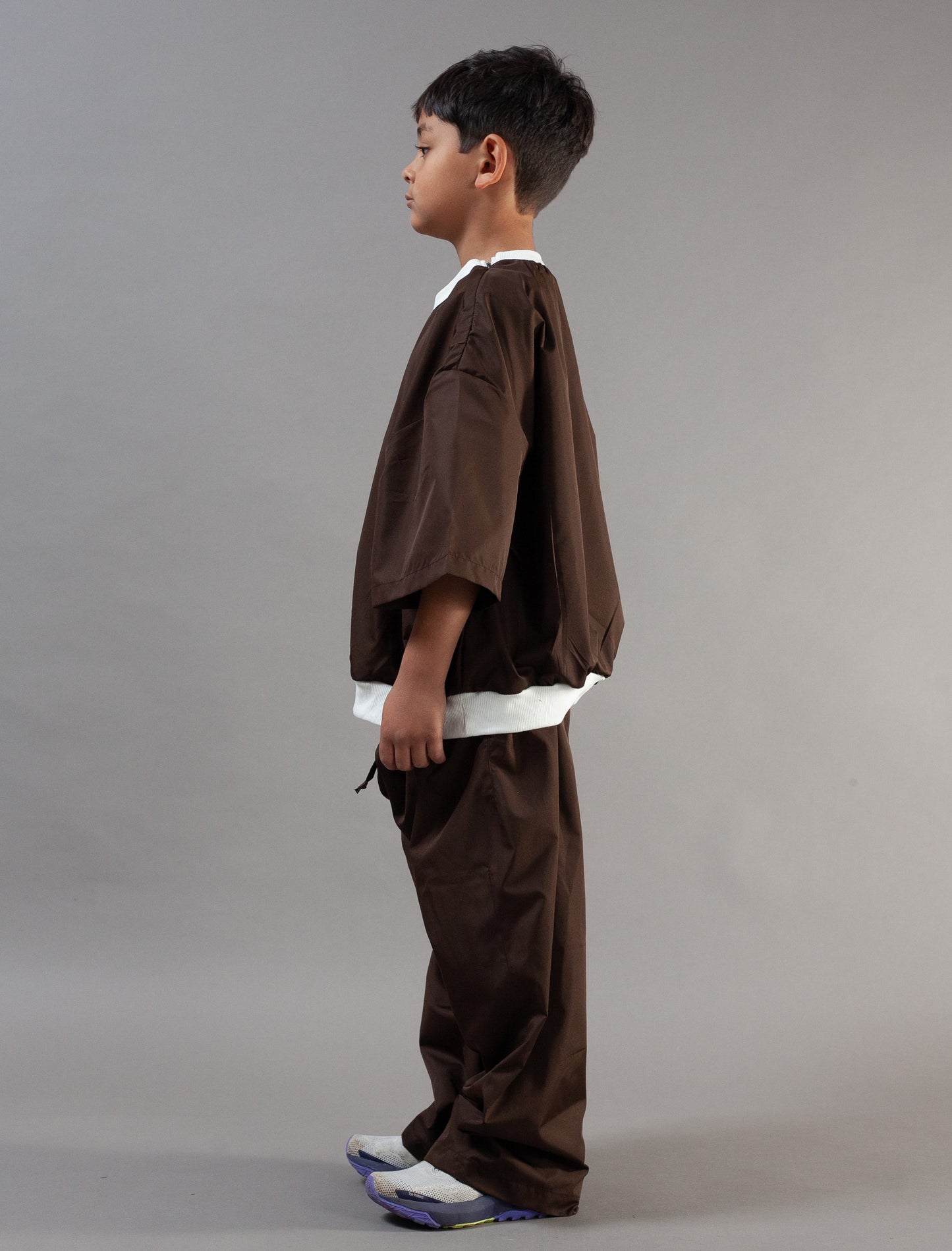 A child model shown in profile wearing a brown two-piece outfit with wide sleeves and matching pants, featuring white contrast trim at the collar and hem, against a plain gray studio background.