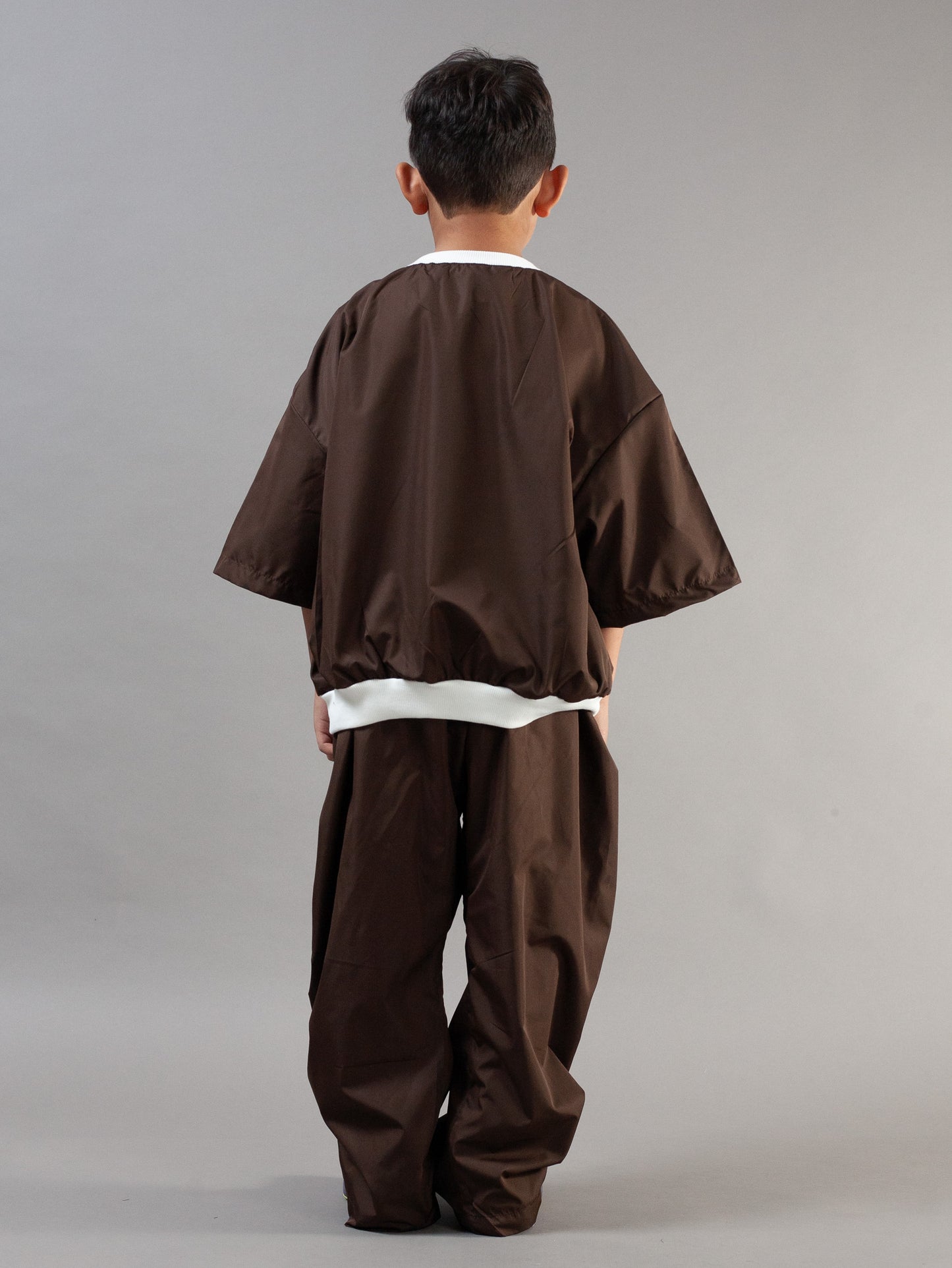 A child viewed from the back wearing a dark brown two-piece outfit with wide sleeves and loose pants, featuring white contrast trim at the collar and waist, against a plain gray studio background.