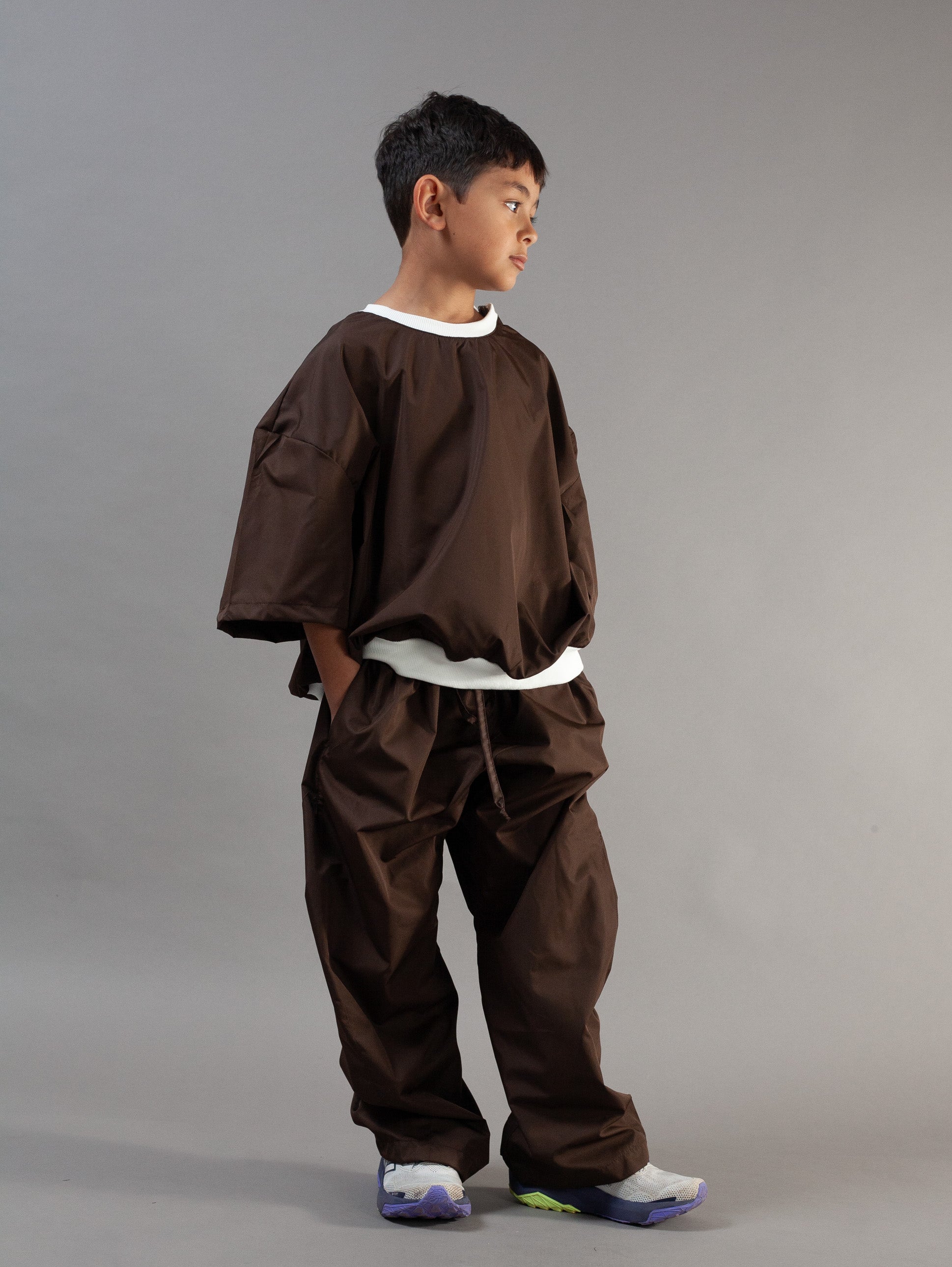 A child model wearing a dark brown two-piece outfit consisting of a loose short-sleeve top and matching pants with white contrast trim, standing against a solid gray studio background.