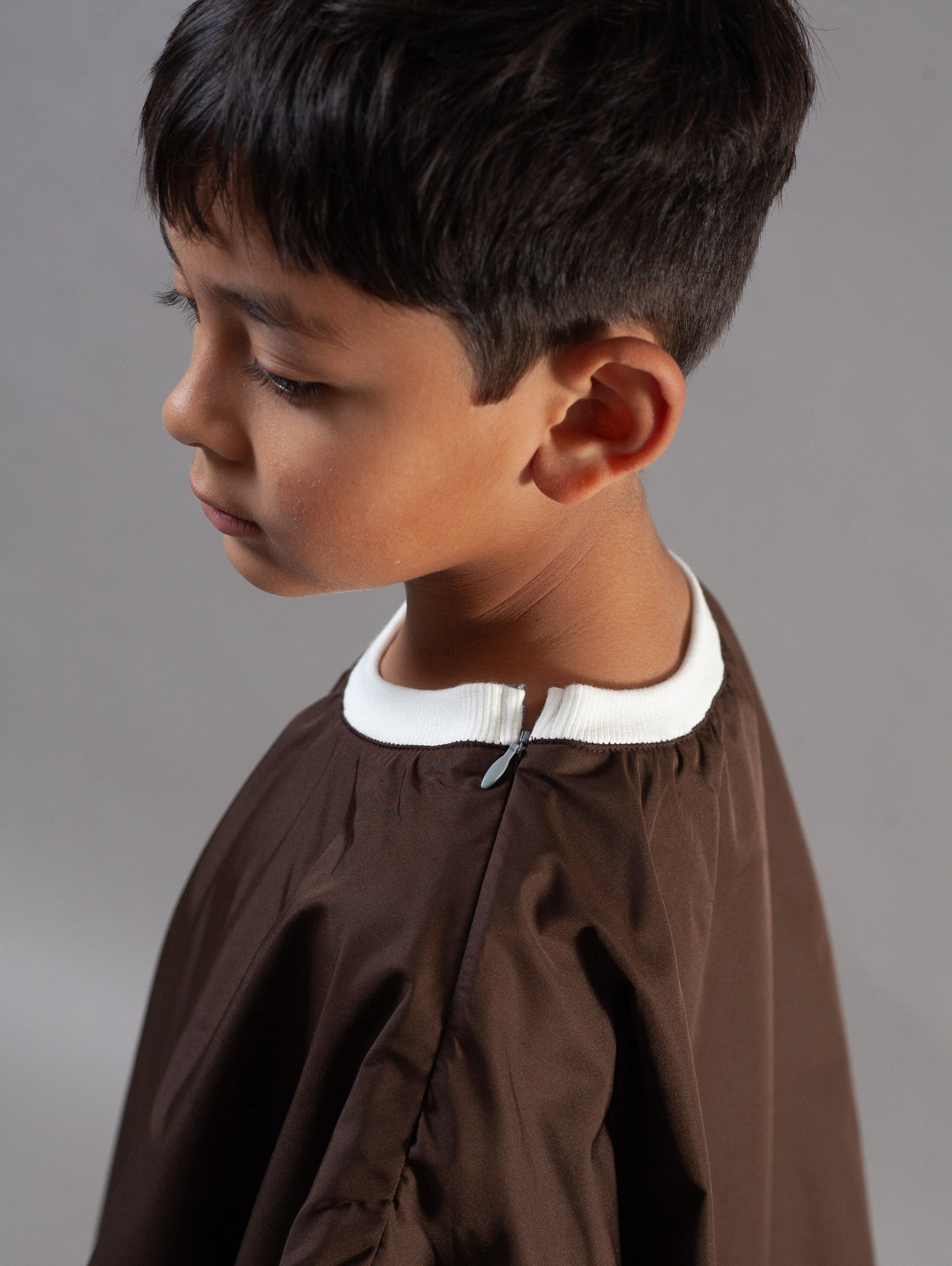 A young boy in profile wearing a brown barber cape with a white collar, photographed against a neutral gray studio background.