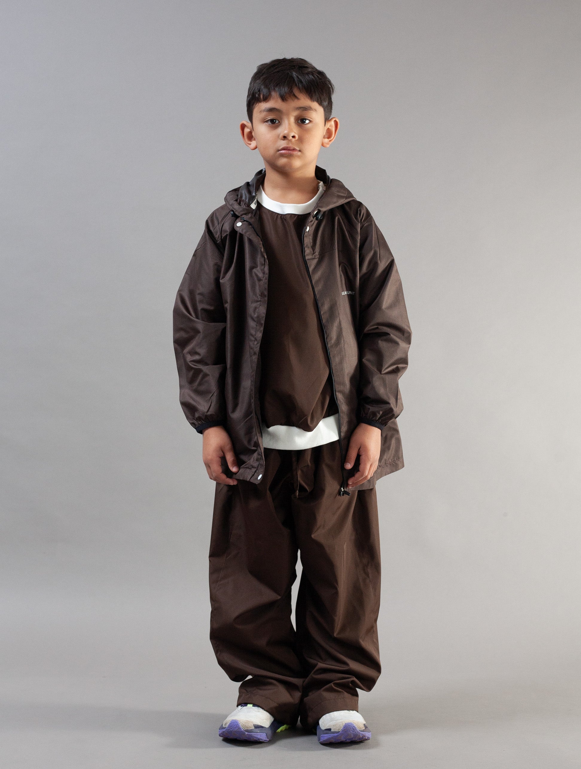 A young boy modeling a dark brown hooded jacket, matching brown sweatshirt, and wide-leg brown pants against a solid gray studio background.