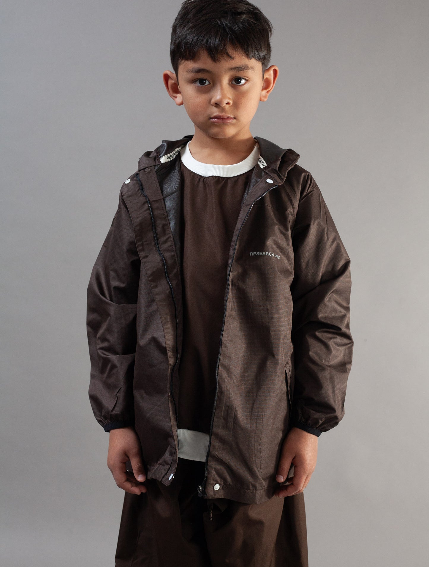 A young boy modeling a dark brown hooded rain jacket over a matching brown t-shirt and brown pants, photographed against a neutral gray studio background.