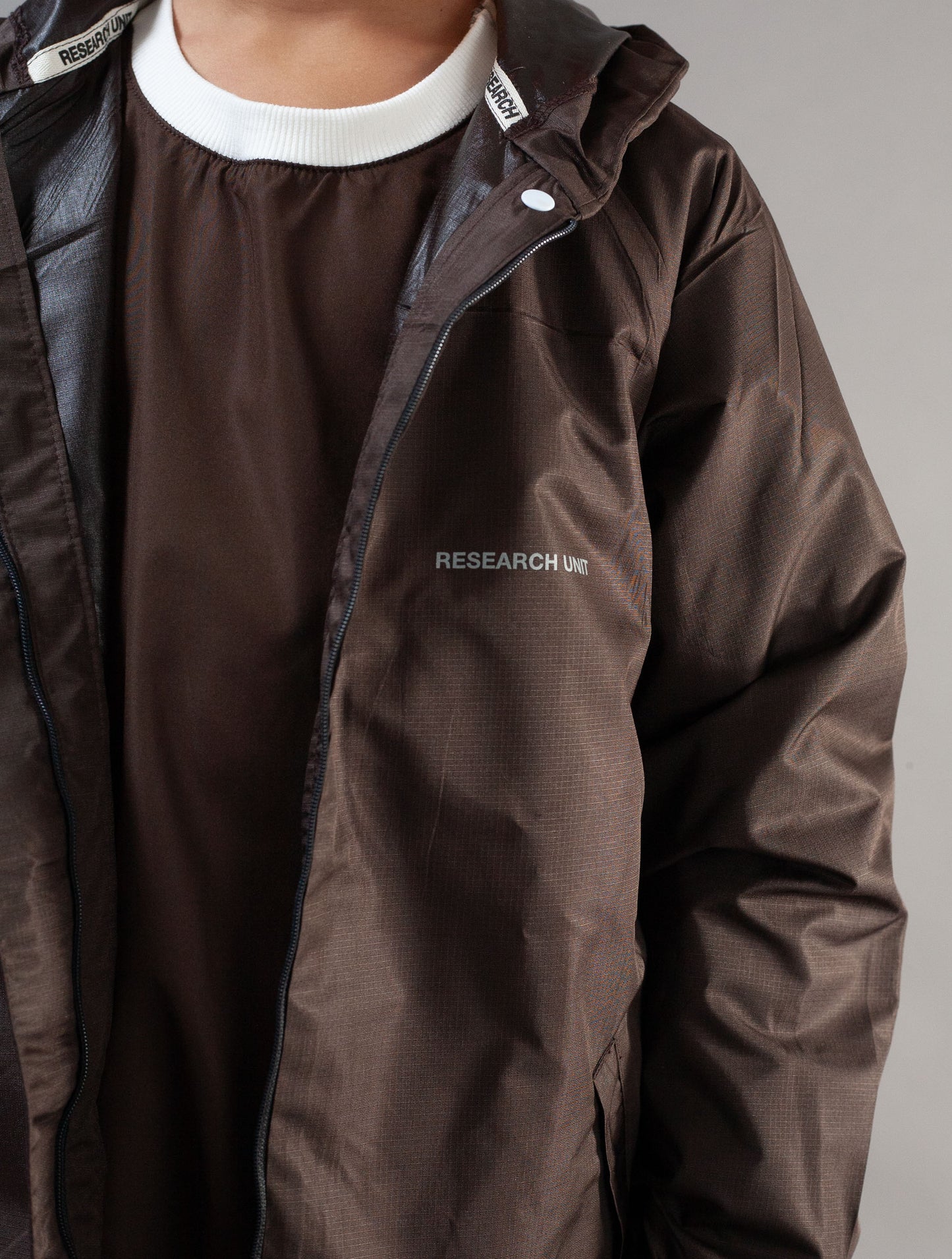 A close-up of a person wearing a dark brown lightweight jacket over a matching brown shirt with a white ribbed collar. The jacket has a small white 'RESEARCH' logo on the left chest and a white snap closure near the collar.