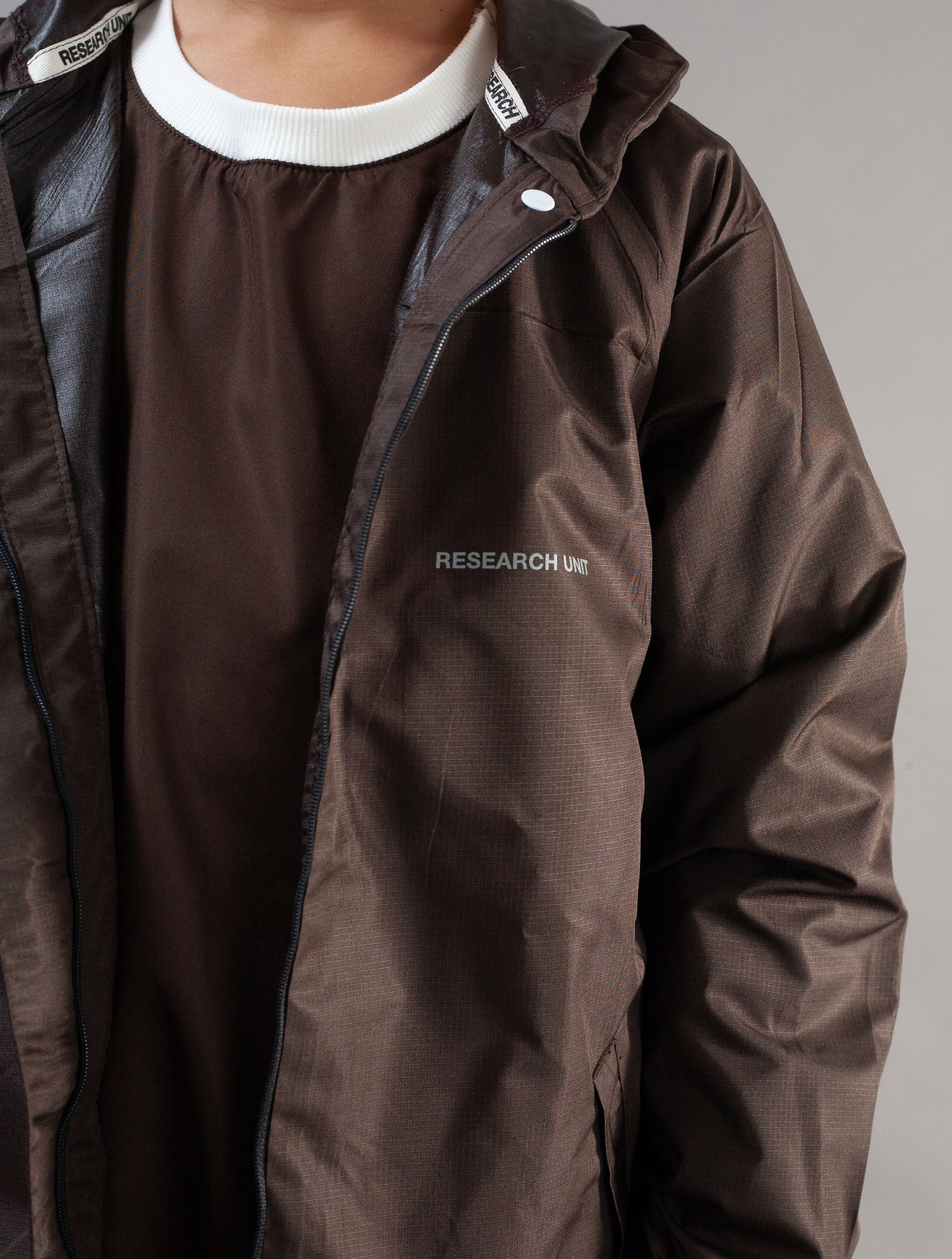 A close-up of a person wearing a dark brown lightweight jacket over a matching brown shirt with a white ribbed collar. The jacket has a small white 'RESEARCH' logo on the left chest and a white snap closure near the collar.
