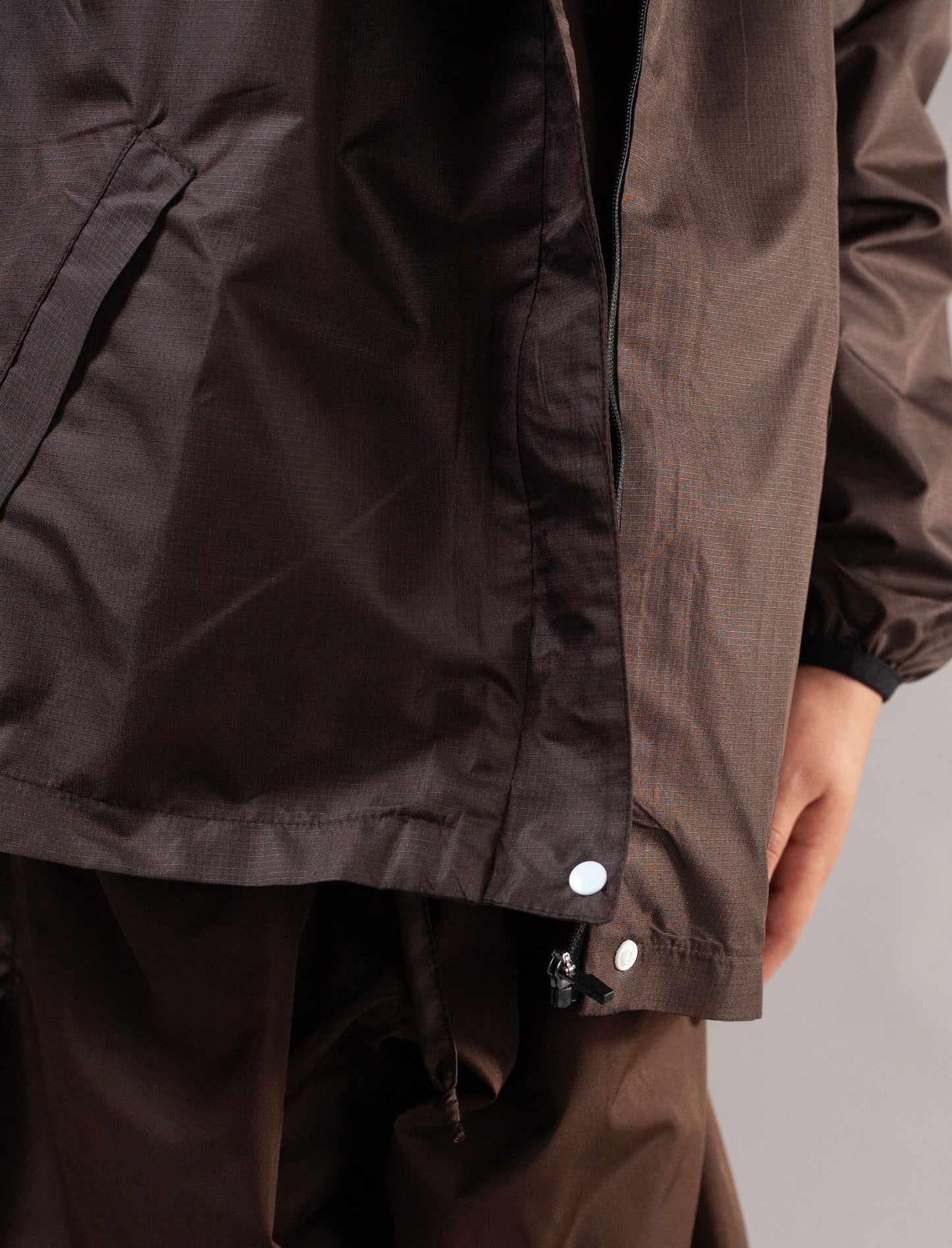 Close-up of a dark brown waterproof jacket with a snap-button closure and matching brown pants, shown on a model against a neutral gray background.
