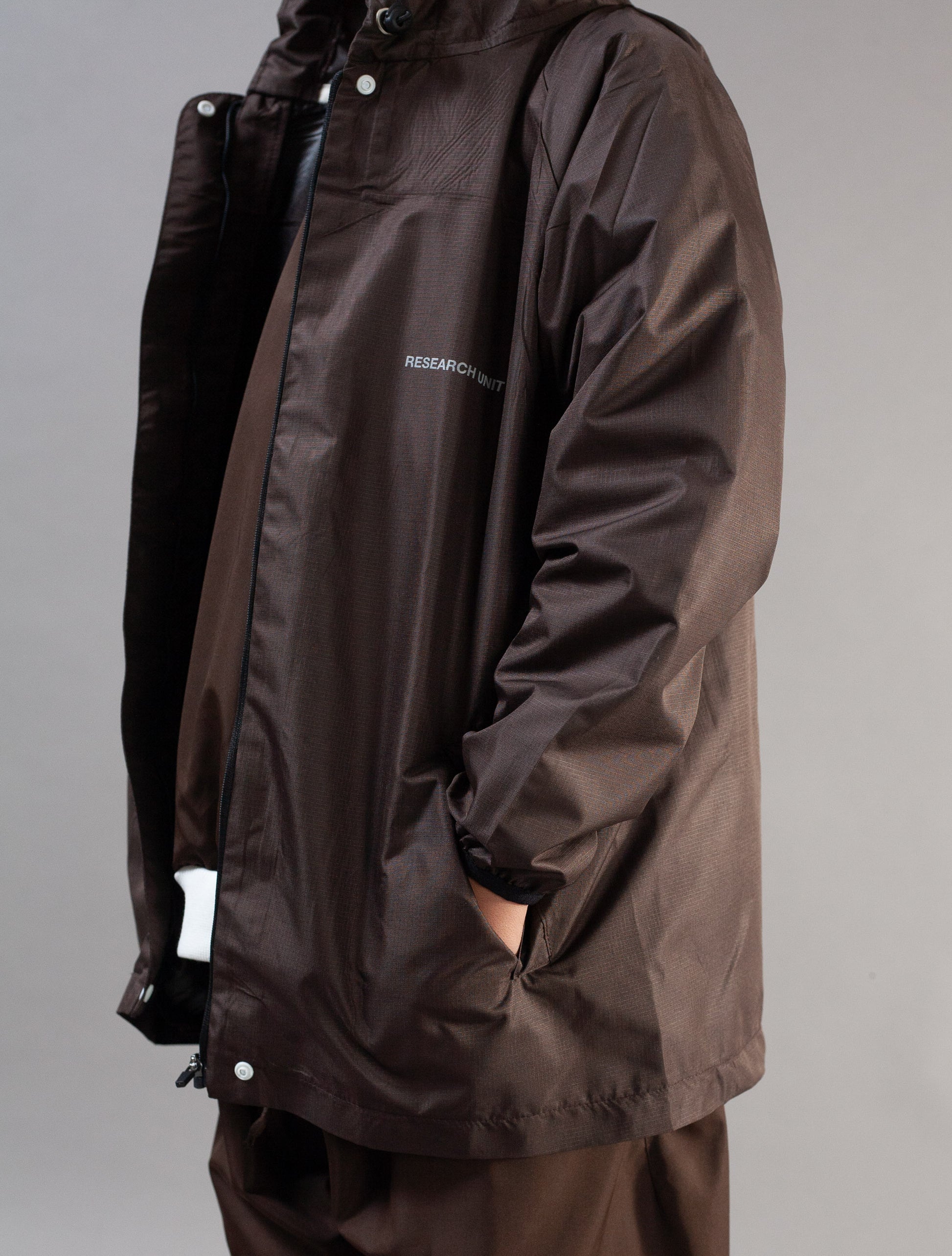 A person wearing a dark brown waterproof jacket with a small white logo on the chest, shown against a plain gray studio background.