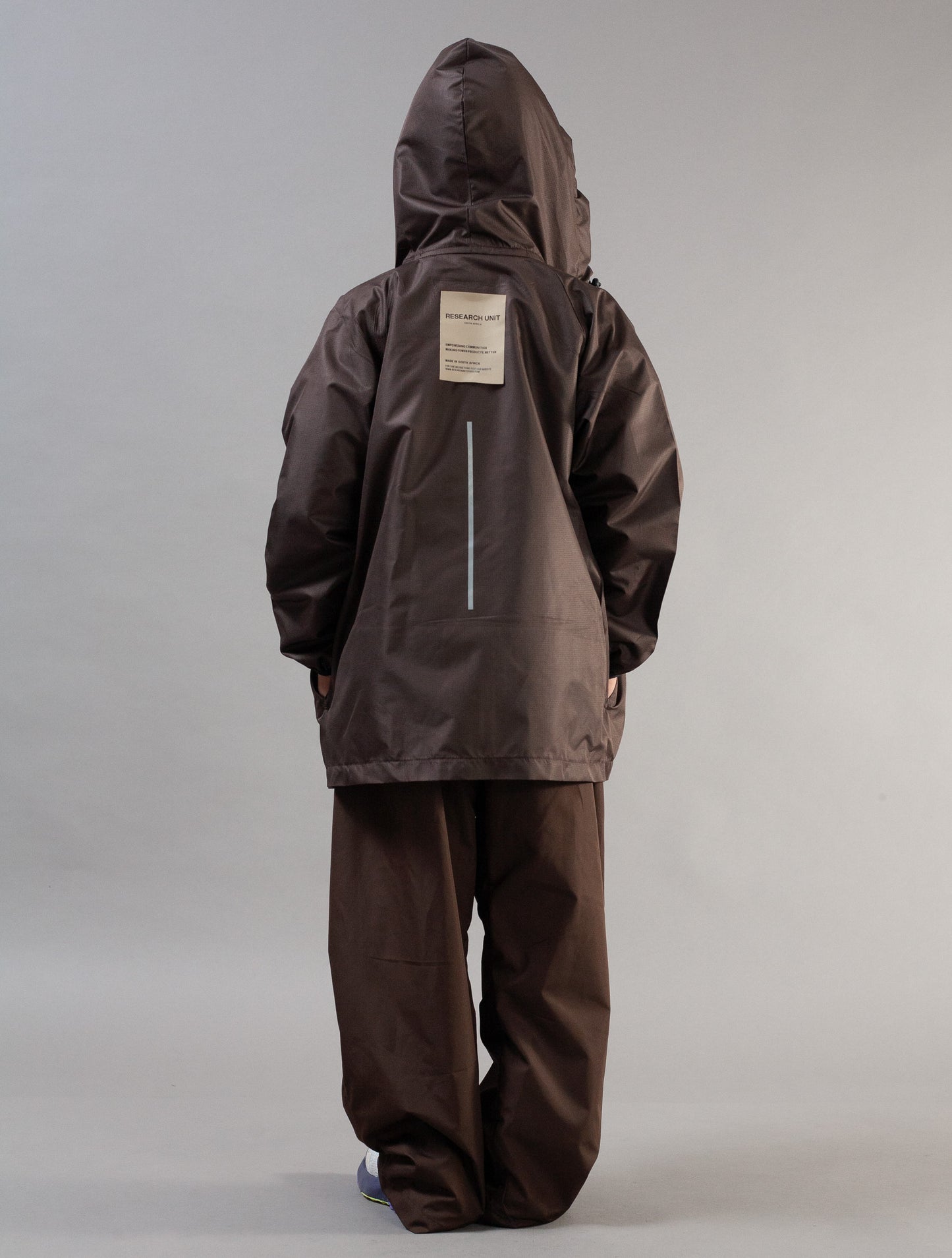 A person shown from the back wearing a dark brown hooded jacket and matching dark brown pants against a plain light gray studio background.