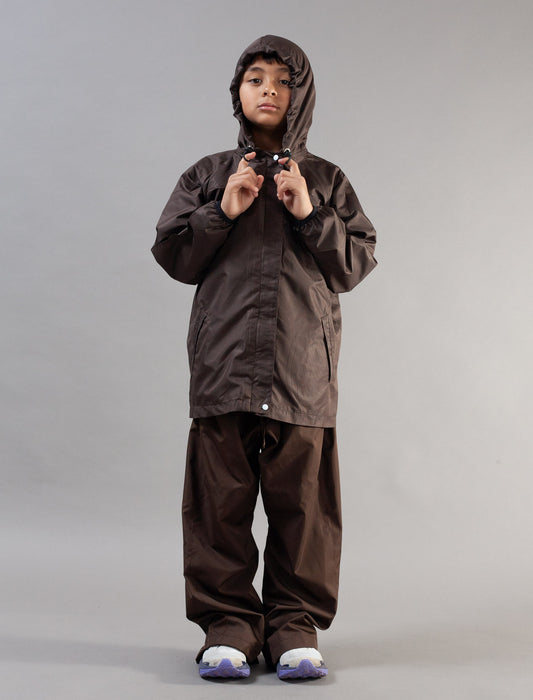 A child wearing a dark brown hooded rain jacket and matching pants, standing against a plain light gray studio background.