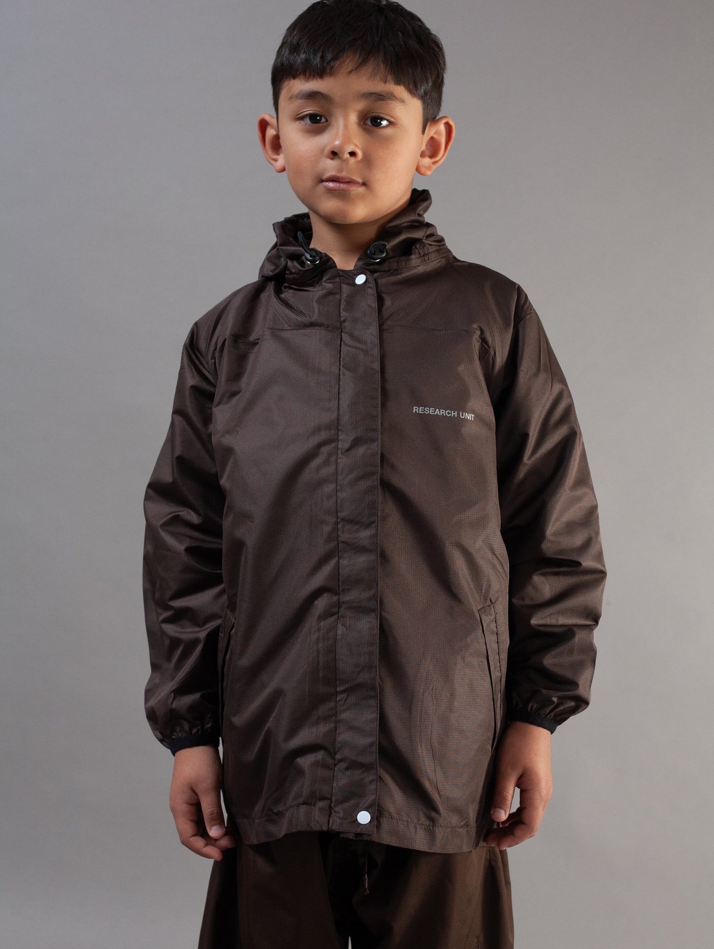 A young boy wearing a dark brown waterproof hooded jacket with a small white brand logo on the left chest, photographed against a neutral gray studio background.