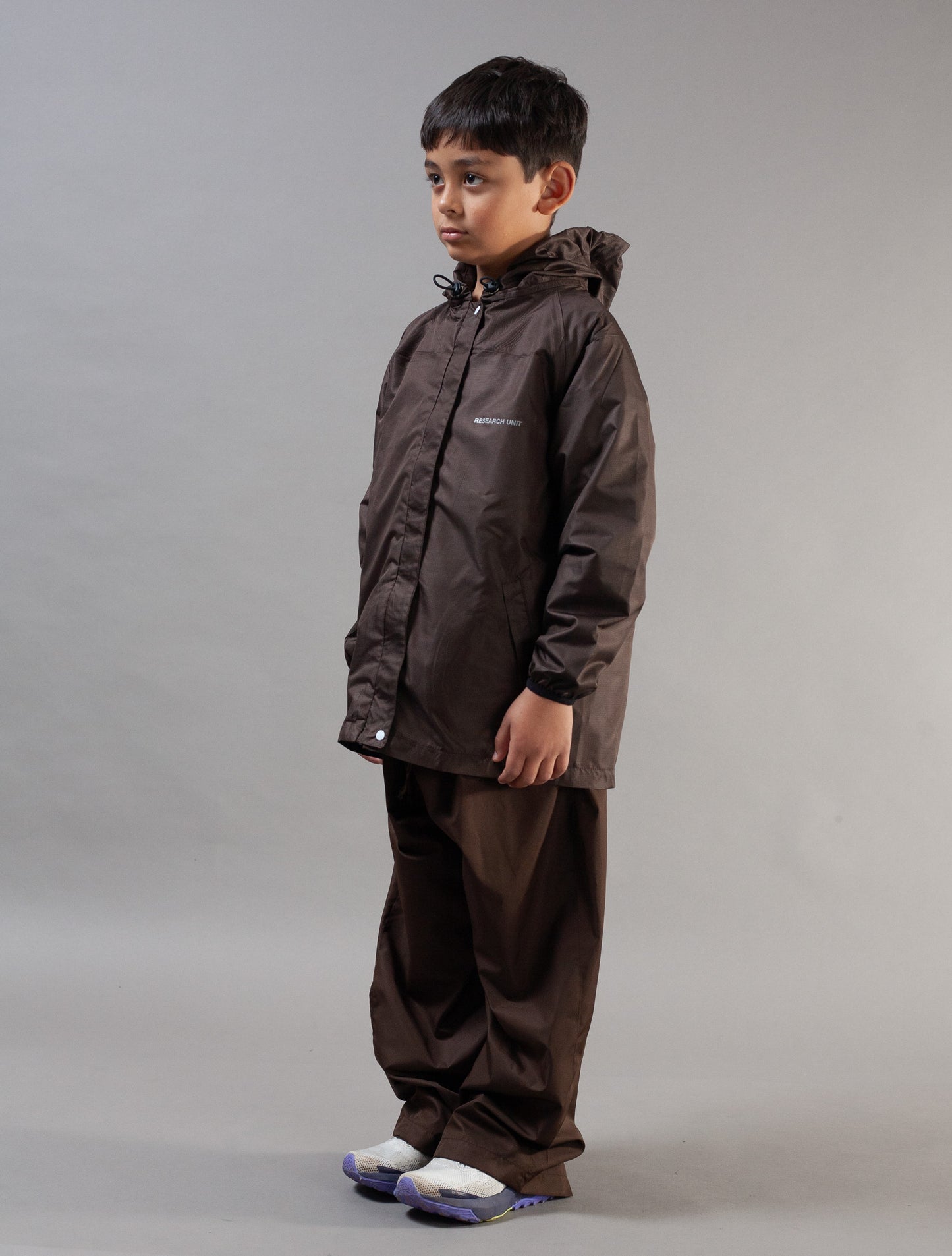 A young boy modeling a dark brown hooded rain jacket and matching brown pants against a neutral gray studio background.