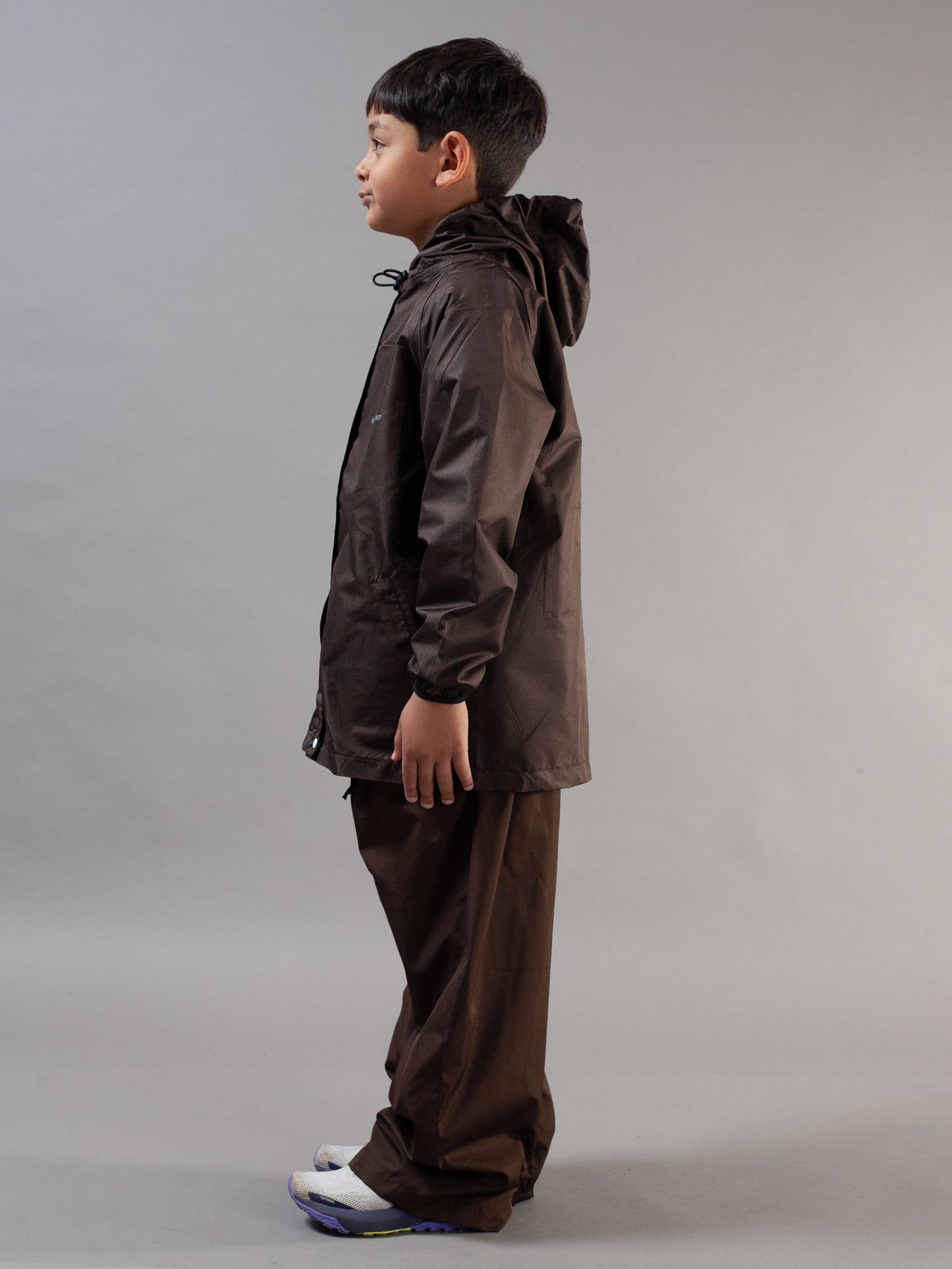 A child model shown in profile wearing a dark brown hooded rain jacket and matching brown pants against a neutral gray studio background.