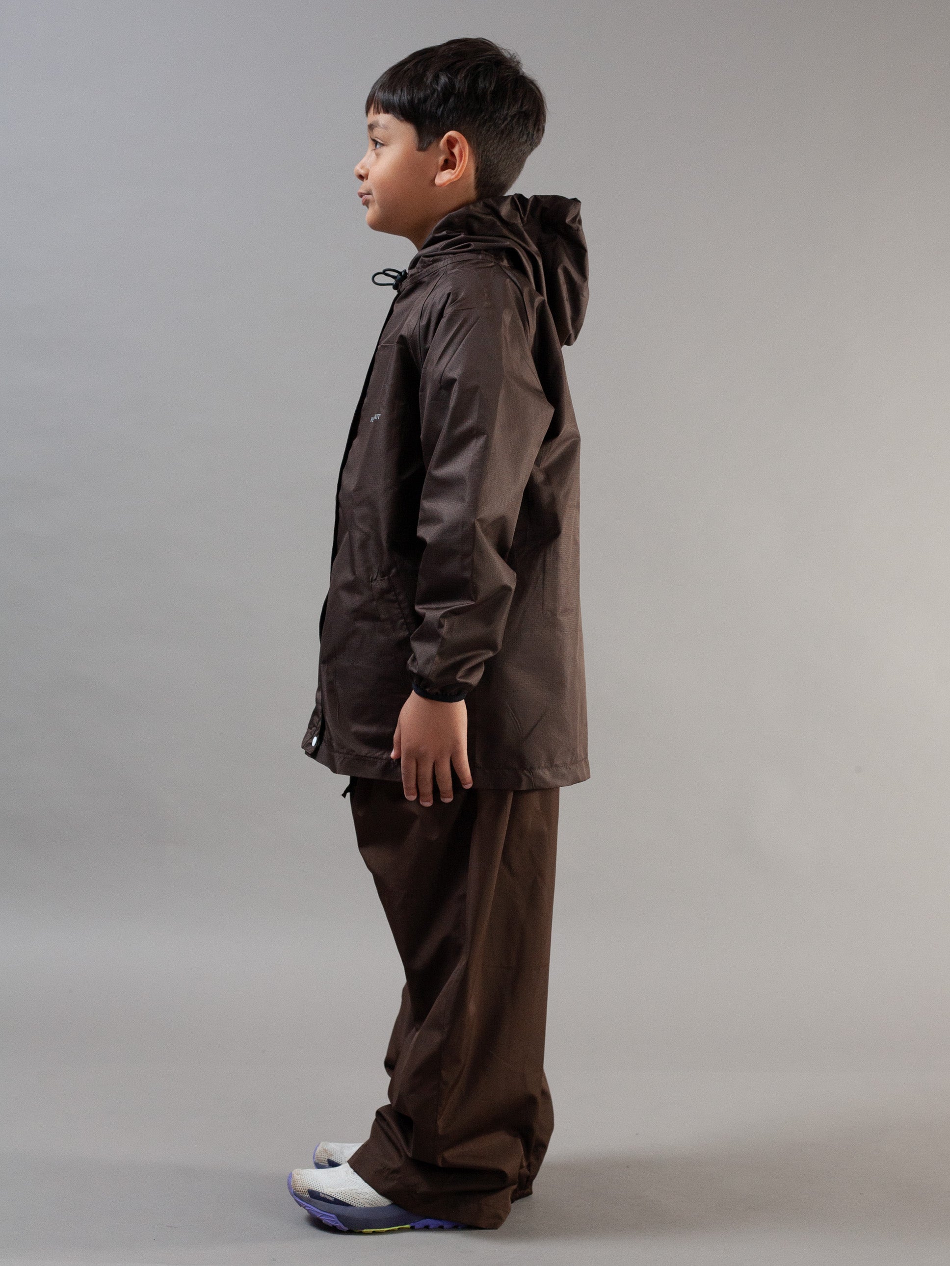 A child model shown in profile wearing a dark brown hooded rain jacket and matching brown pants against a neutral gray studio background.