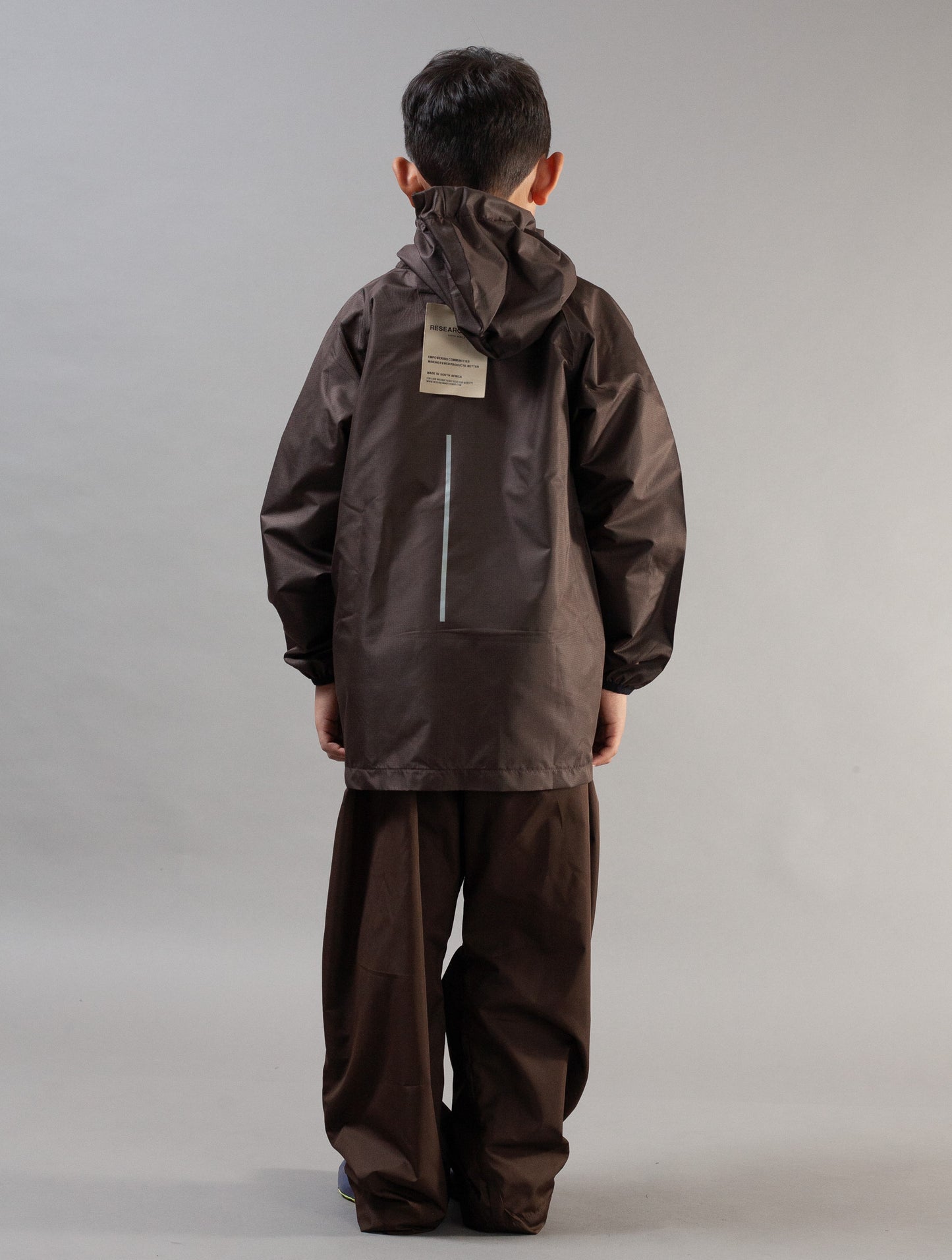 A child viewed from the back wearing a dark brown hooded rain jacket and matching dark brown pants against a plain light gray studio background.