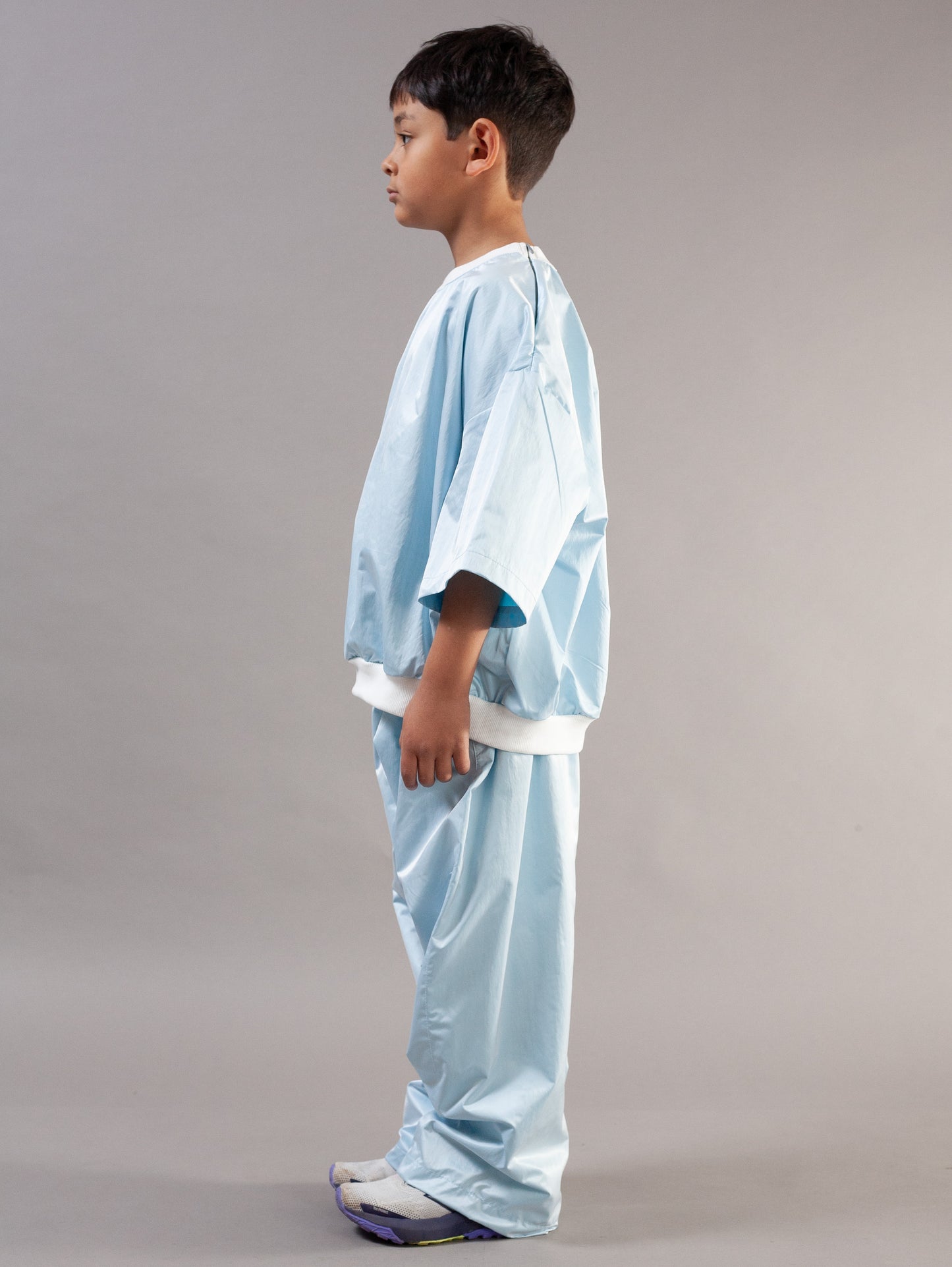 A young boy modeling a light blue kids set, consisting of a short-sleeve top and matching pants, standing against a neutral gray studio background.
