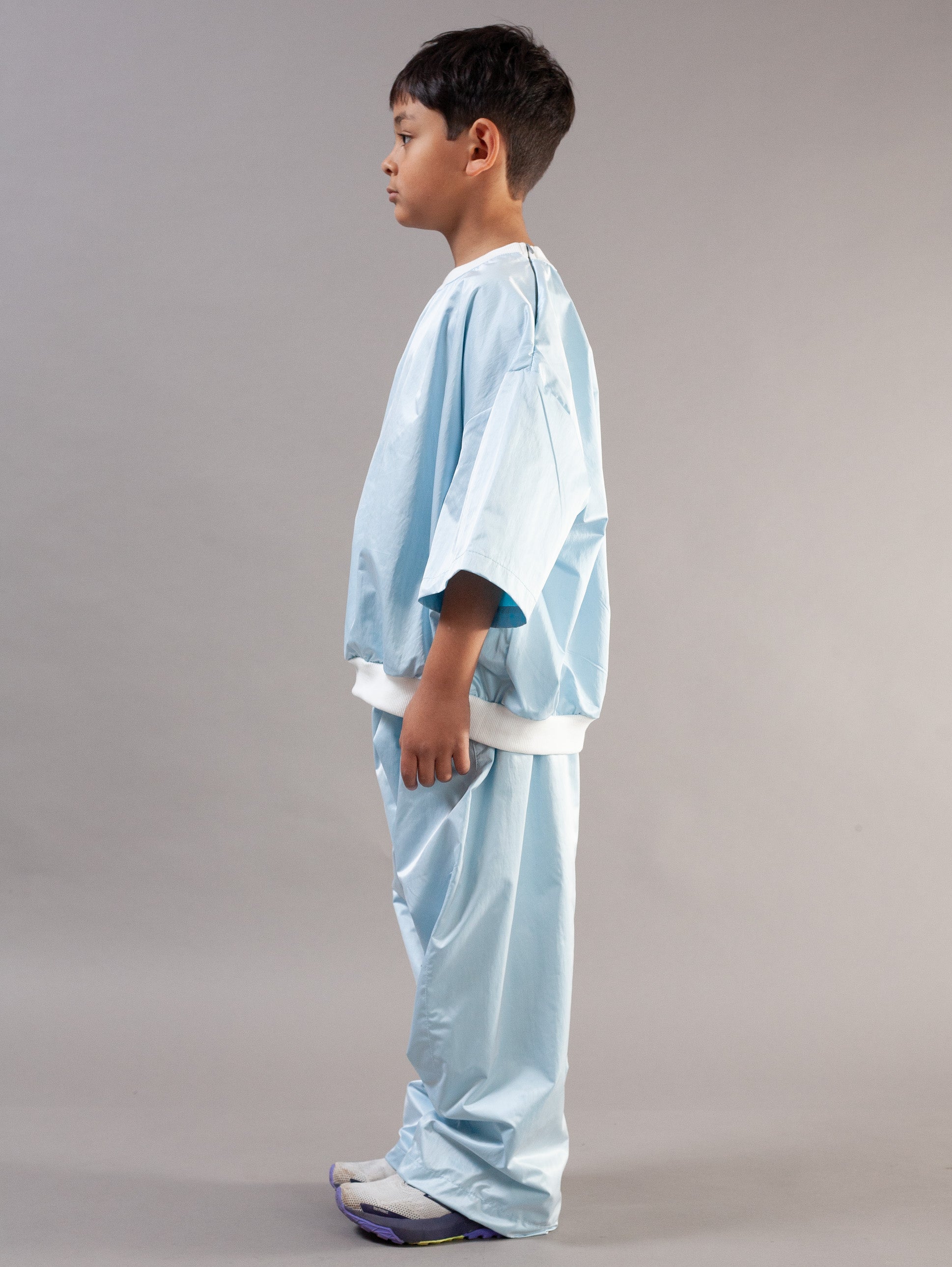 A young boy modeling a light blue kids set, consisting of a short-sleeve top and matching pants, standing against a neutral gray studio background.