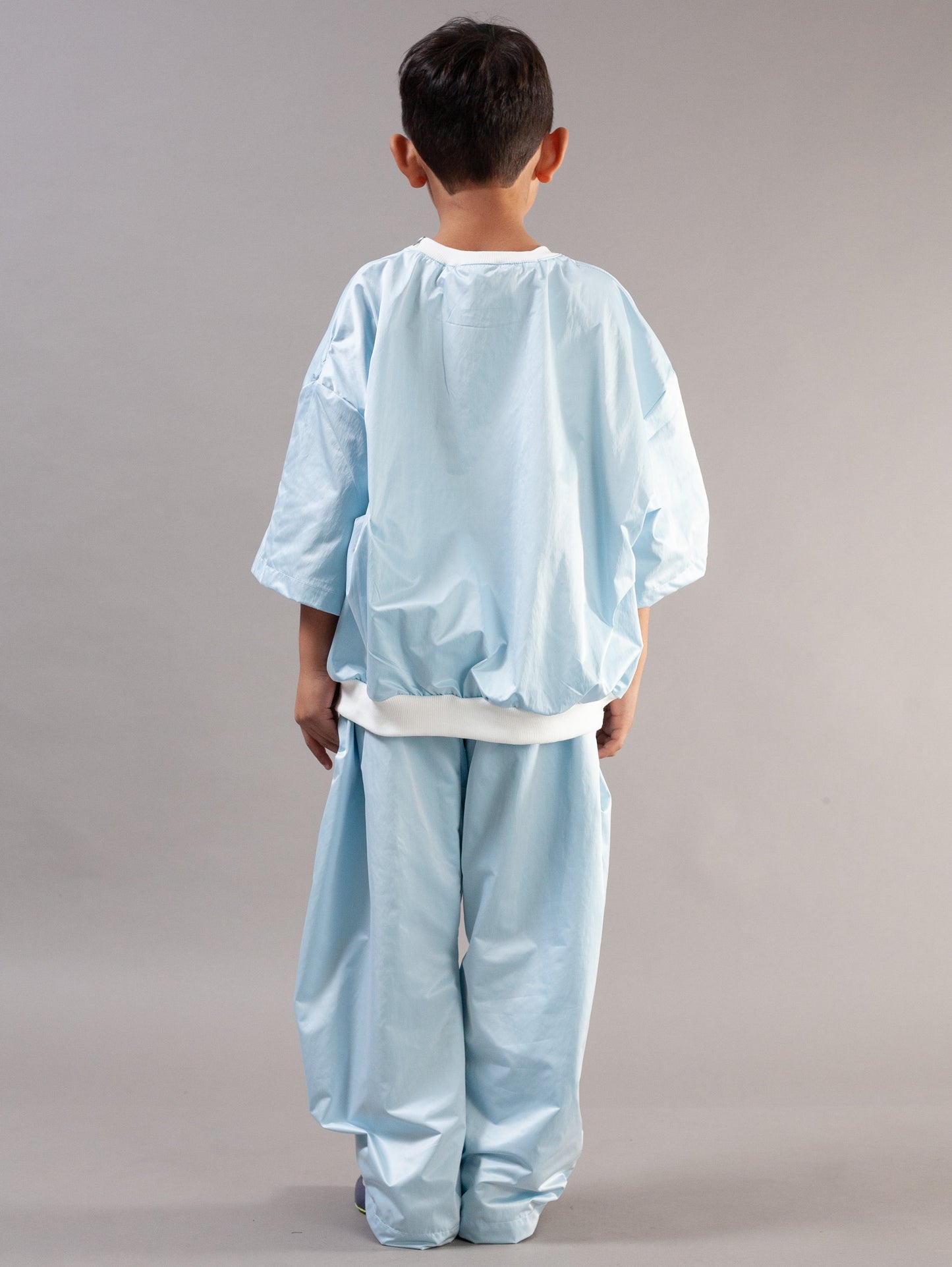 A young boy modeling a light blue kids set, consisting of a short-sleeve top and matching pants, standing against a neutral gray studio background.
