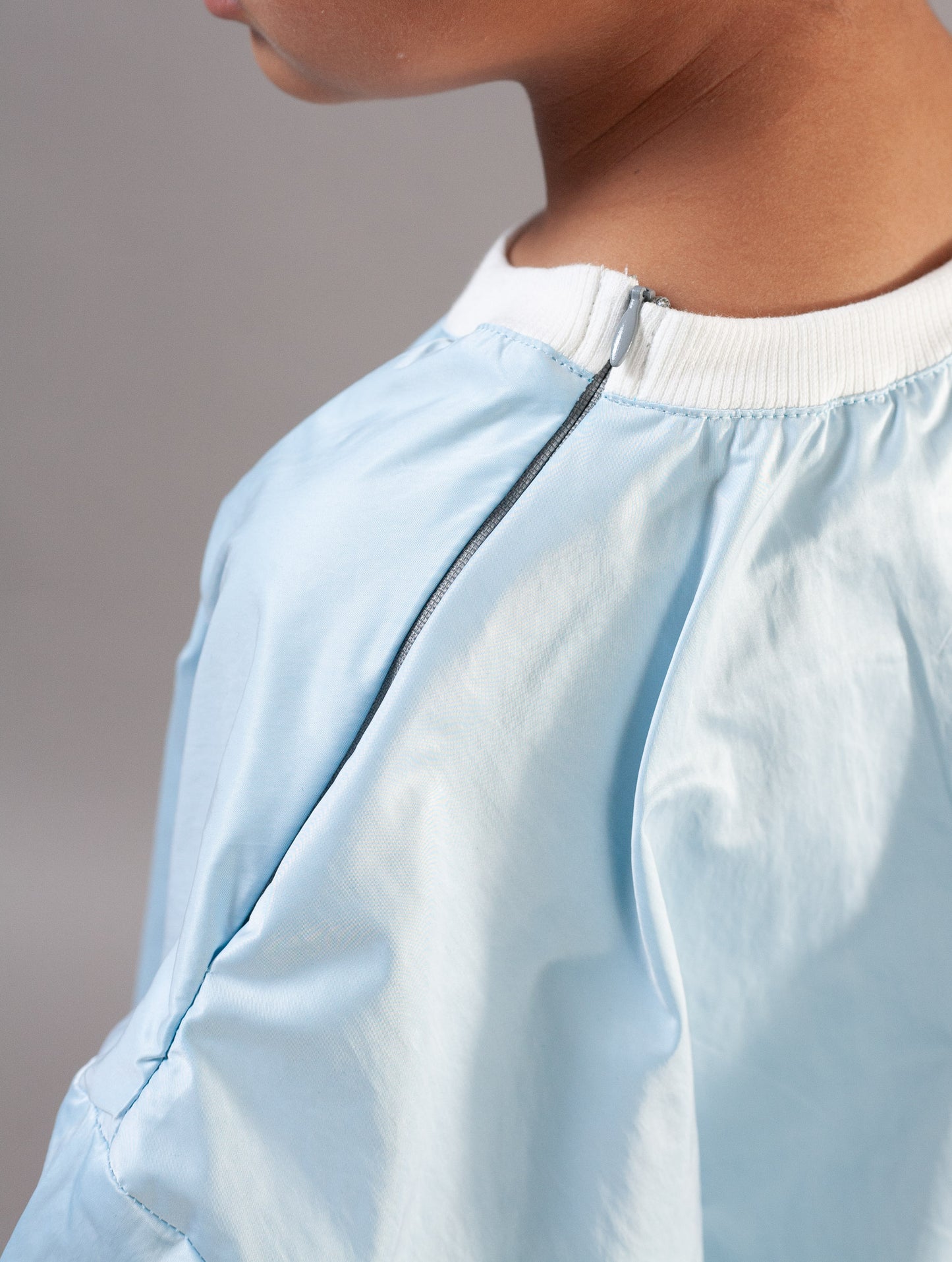 Close-up of a person wearing a light blue zippered garment with a white ribbed collar, cropped from the side against a plain gray background.