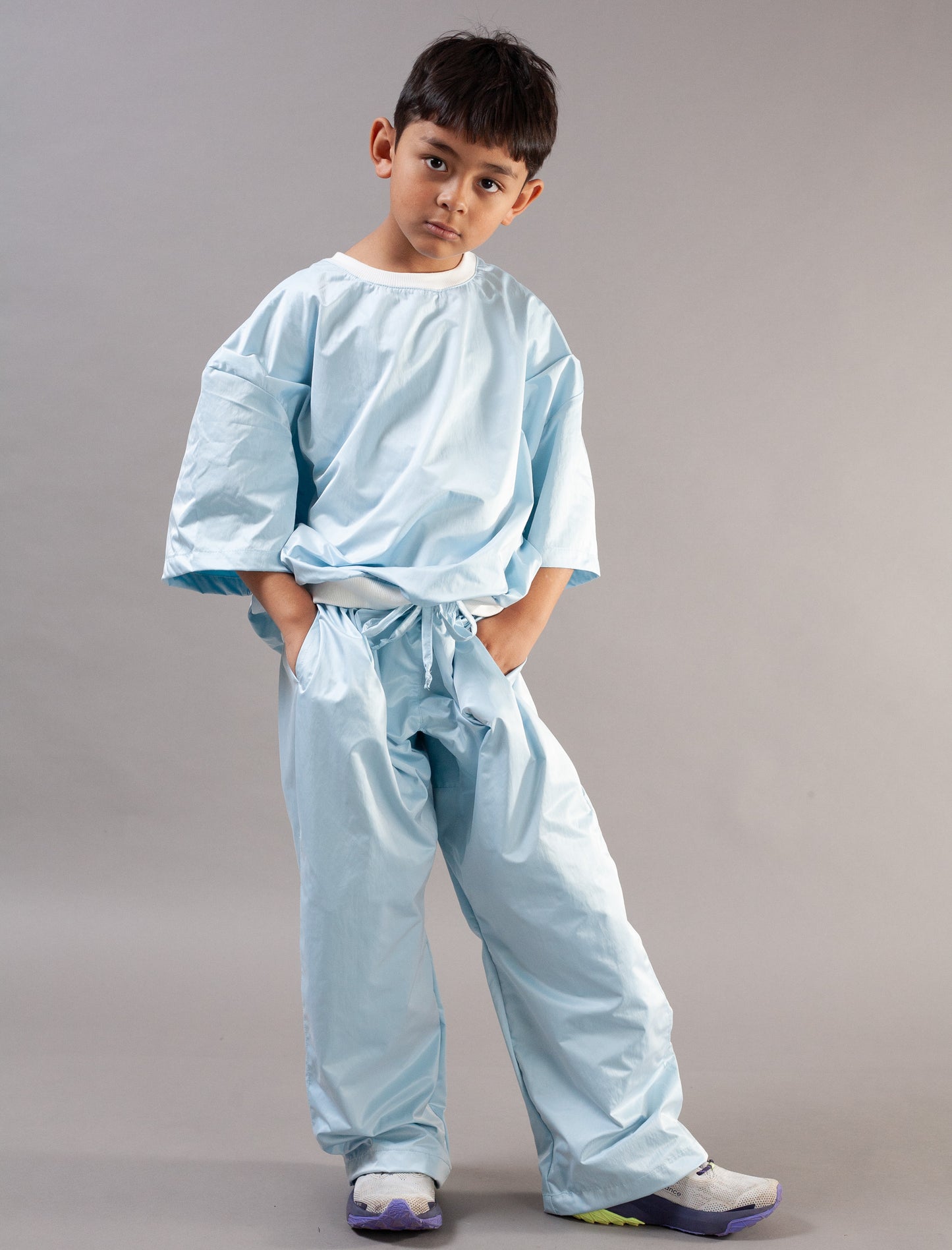 A young boy modeling a light blue two-piece set or training uniform consisting of a short-sleeve top and matching pants, standing against a solid gray studio background.