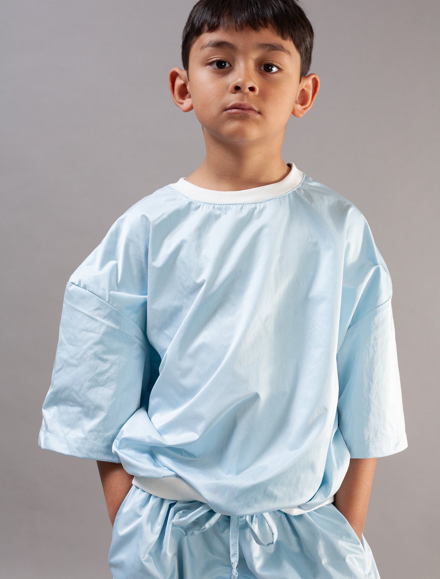 A young boy wearing a light blue set with elbow-length sleeves and a matching drawstring waist, photographed against a plain gray studio background.