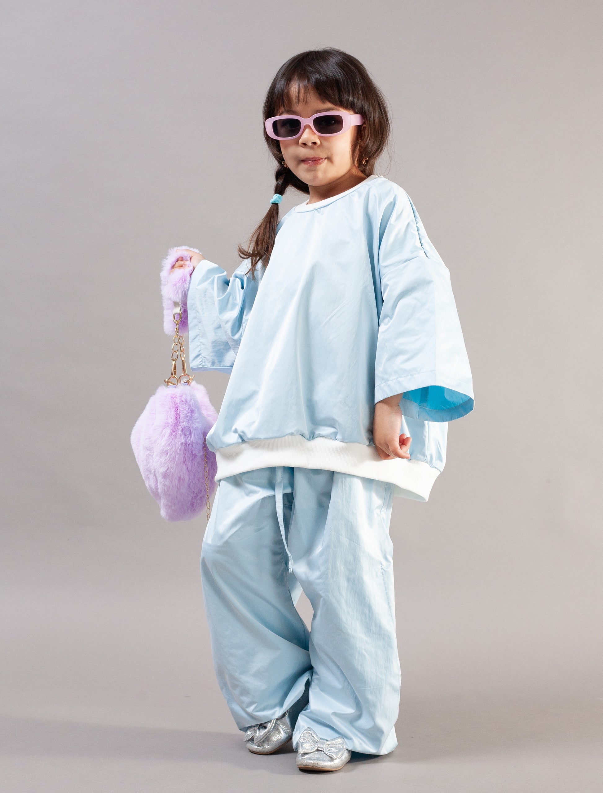 A young child wearing a light blue oversized two-piece outfit with wide sleeves and pants, pink sunglasses, and holding a fluffy purple bag against a plain gray studio background.