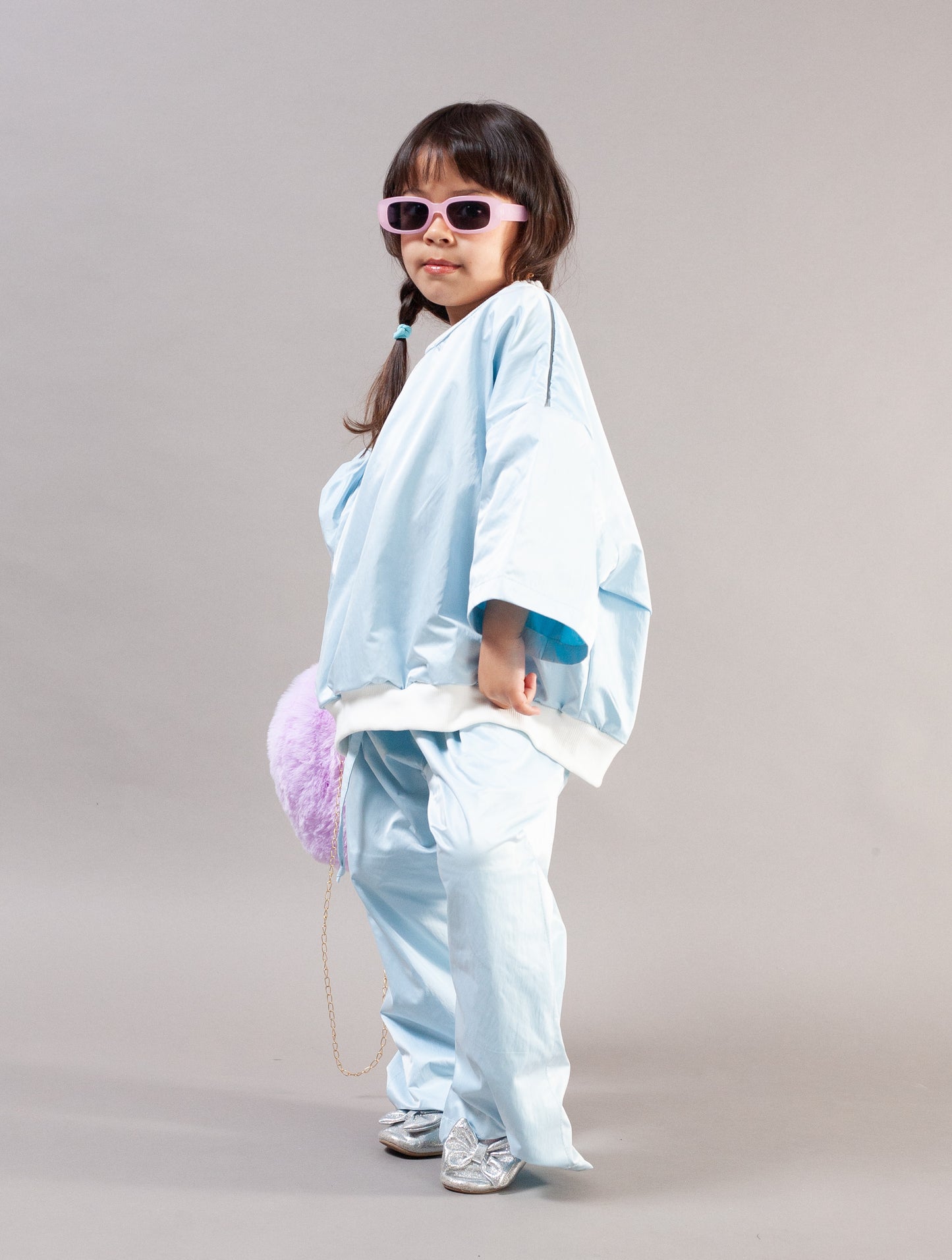 A young girl modeling a light blue kimono-style outfit with matching pants, pink sunglasses, and a fuzzy lavender bag against a plain gray studio background.