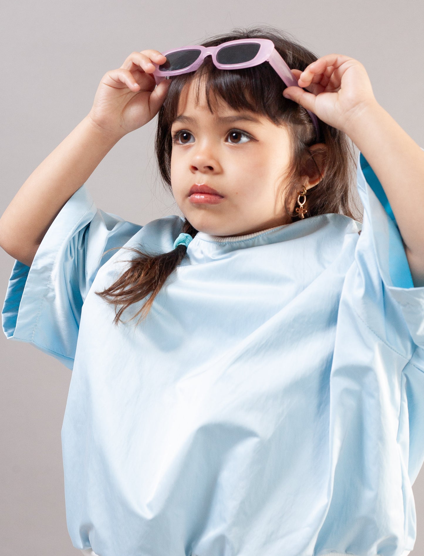 A young girl with dark hair wearing a light blue satin-like top and pink sunglasses, posing with her hands on her head against a solid gray background.