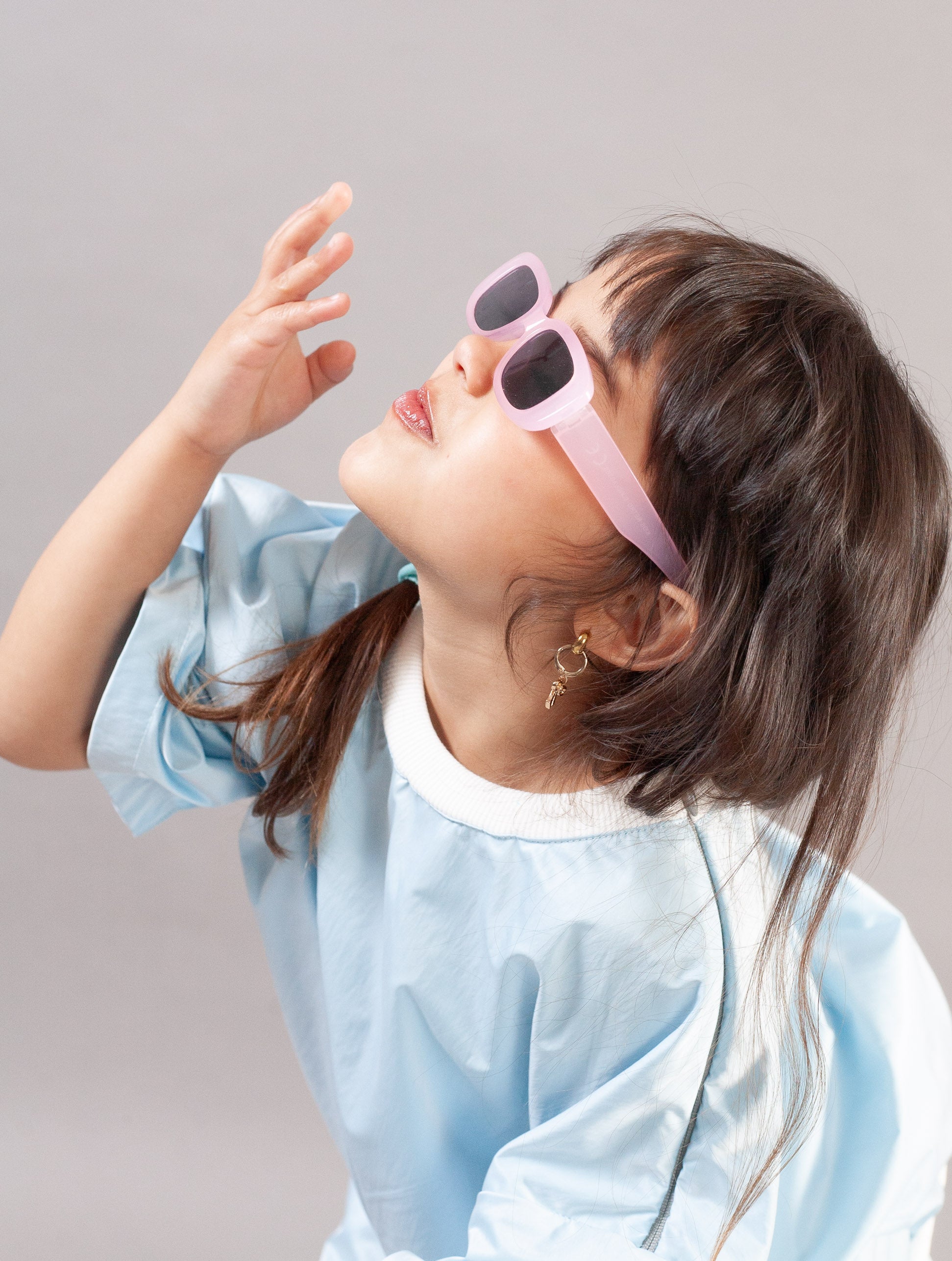 A young girl with dark brown hair wearing light pink square sunglasses and a light blue painter's smock over a white shirt, looking upwards against a solid gray background.