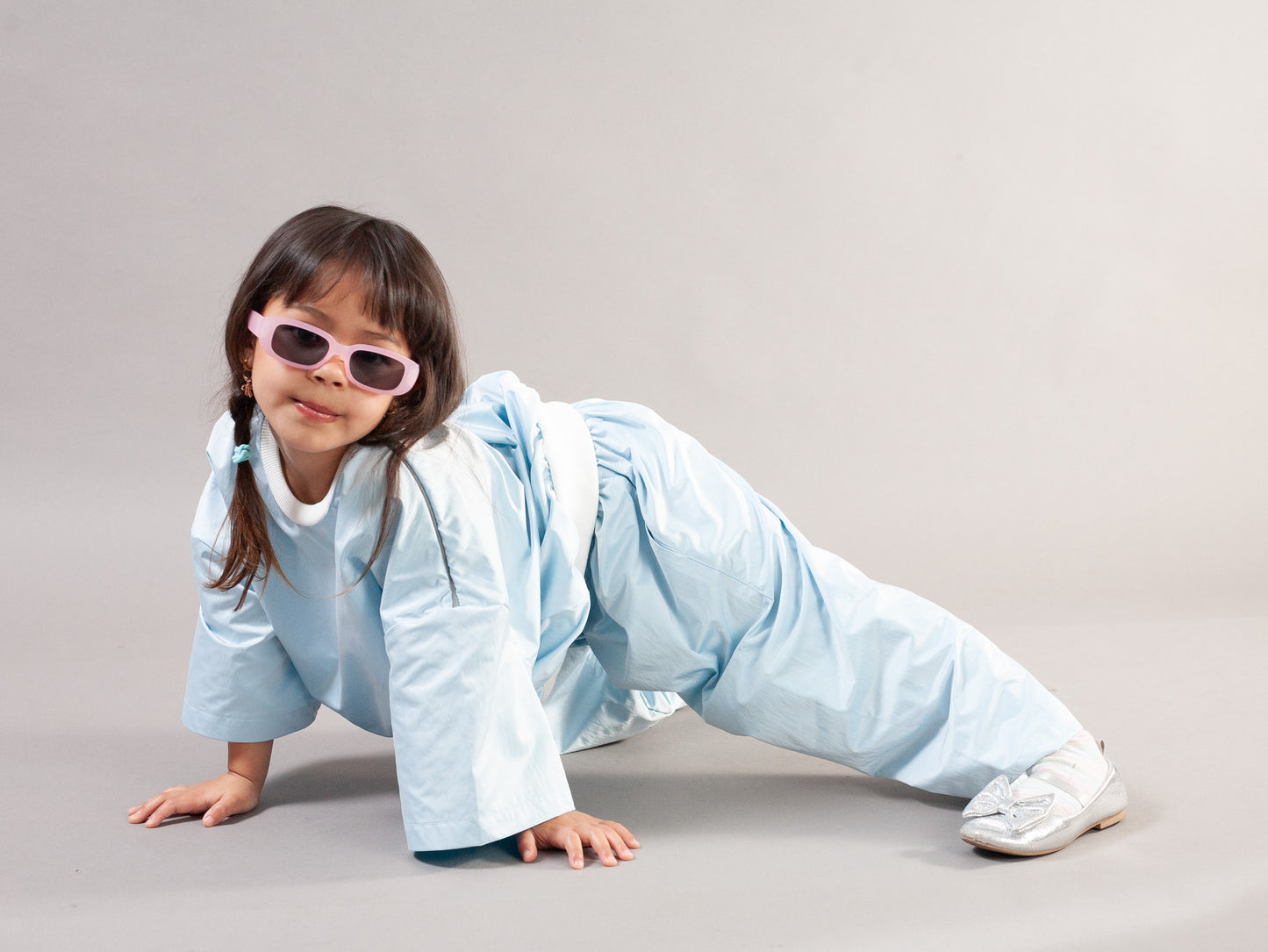 A young girl in a light blue martial arts uniform and white sneakers poses on her hands and knees on a light gray studio background, wearing pink-framed sunglasses.