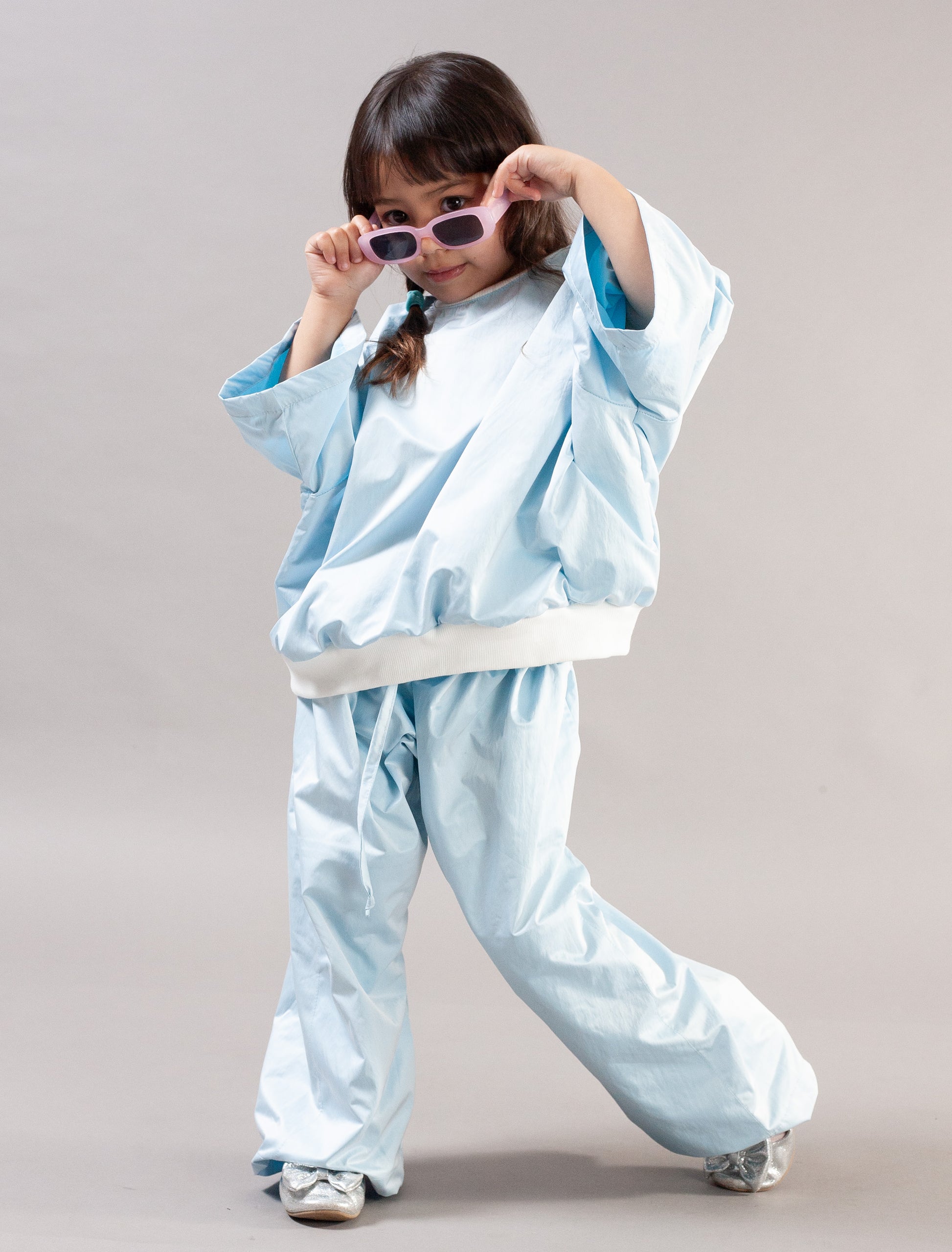 A young child modeling a light blue two-piece tracksuit with a white waistband, styled with pink sunglasses and white sneakers against a neutral studio background.