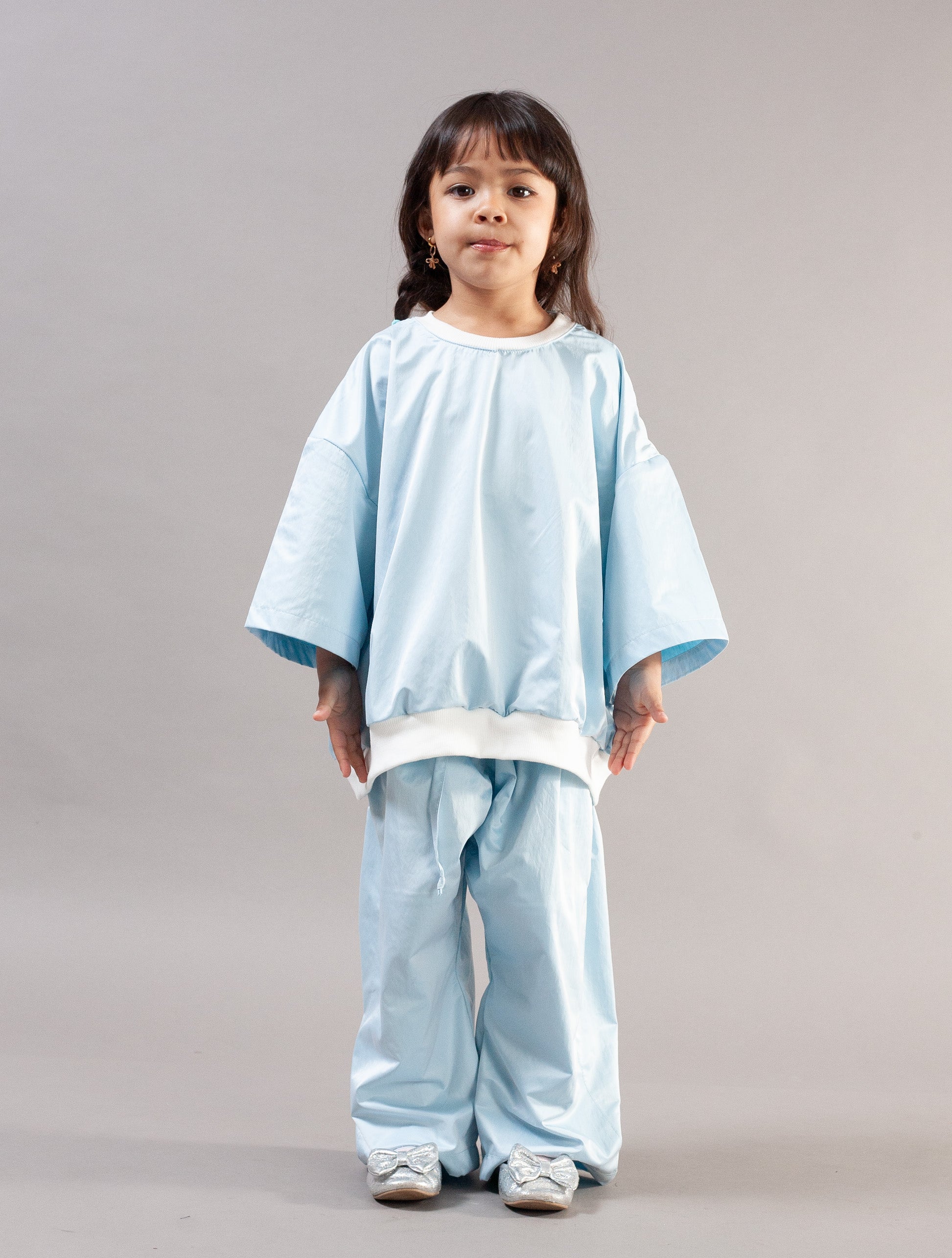 A young girl with dark hair in a braid stands against a neutral gray studio background, wearing a light blue two-piece outfit with wide sleeves and matching pants, featuring white contrast trim at the collar and hem.