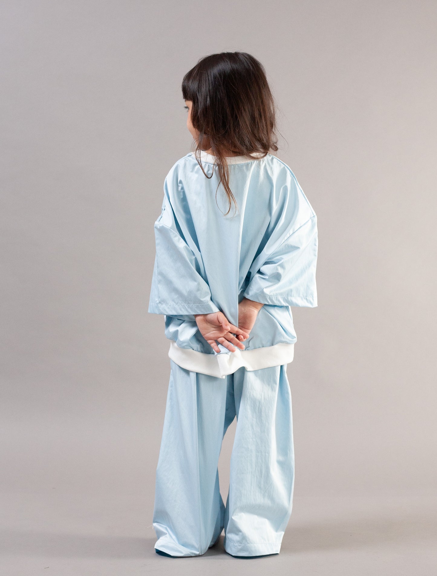 A child standing with their back to the camera, wearing a light blue kimono-style outfit with wide sleeves and matching pants, featuring a white waistband detail, set against a plain light gray studio background.