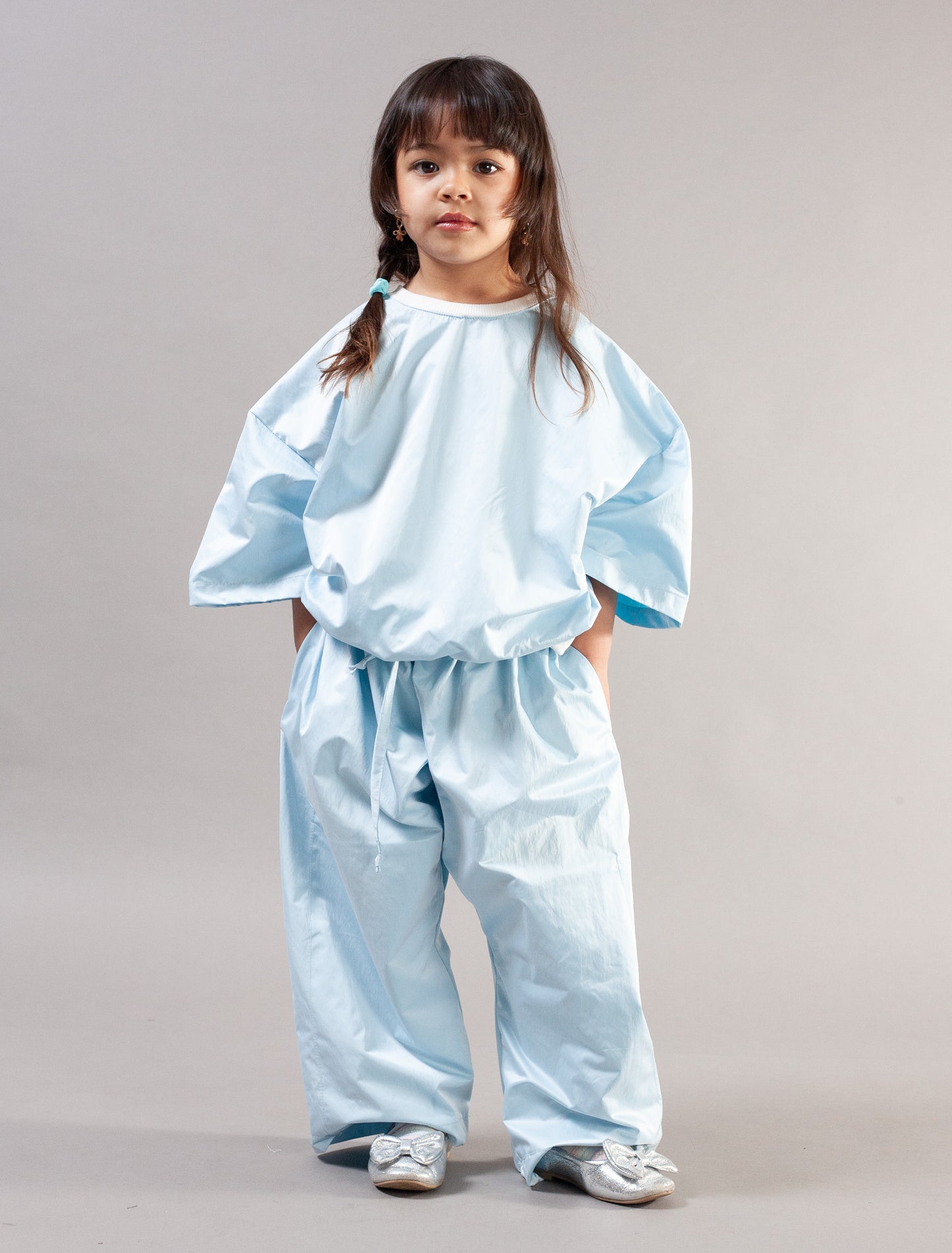 A young girl with dark hair in a braid stands wearing a light blue two-piece outfit consisting of a loose short-sleeve top and matching wide-leg pants, posed against a neutral gray studio background.