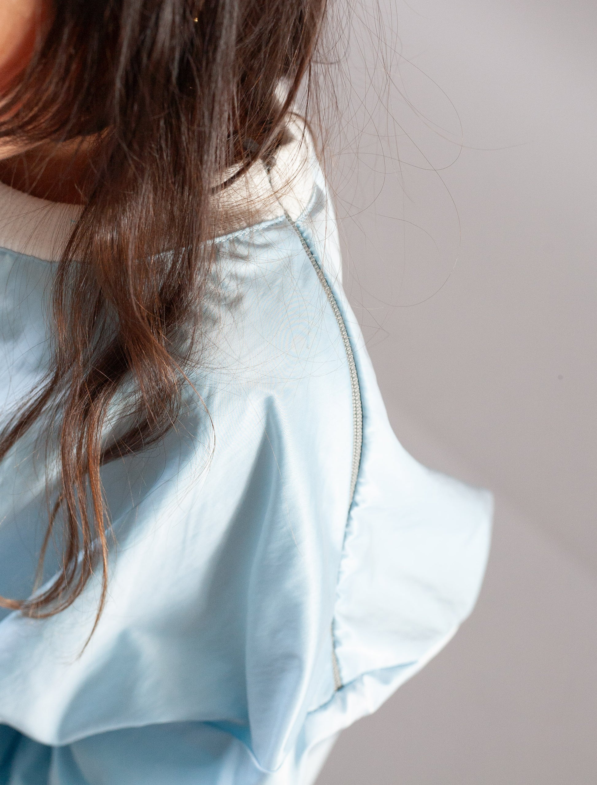 Close-up of a person wearing a light blue ruffled blouse or dress, showing the back and shoulder area with long brown hair against a plain light gray background.