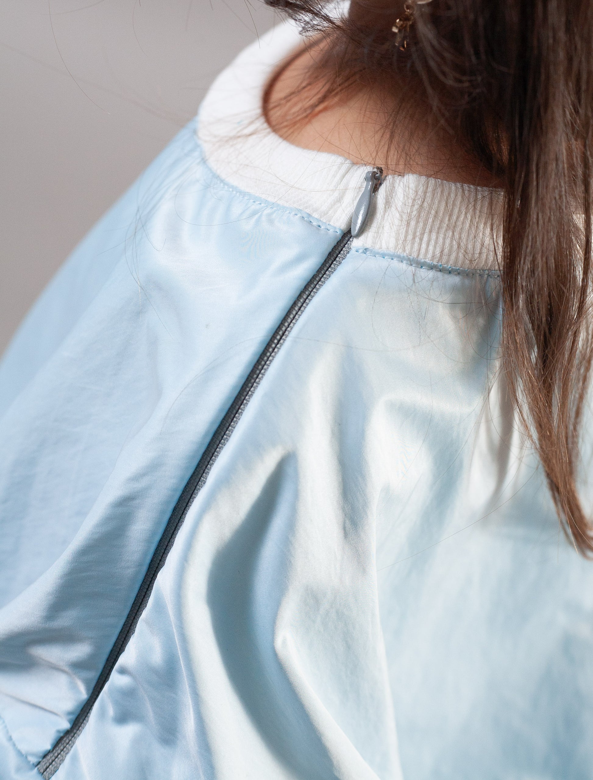 Close-up of a light blue satin bomber jacket with a white ribbed knit collar, featuring a silver zipper and a model's brown hair visible.