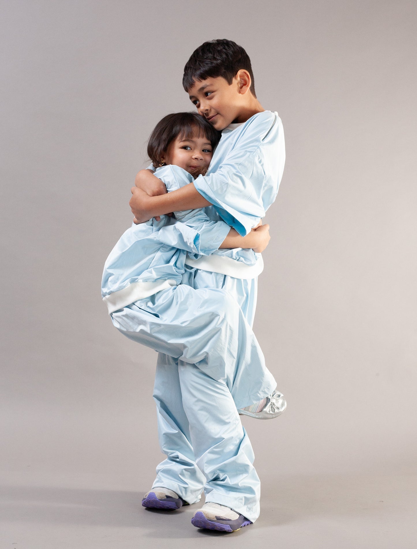 Two children wearing matching light blue martial arts uniforms with white belts, posed against a plain light gray studio background.