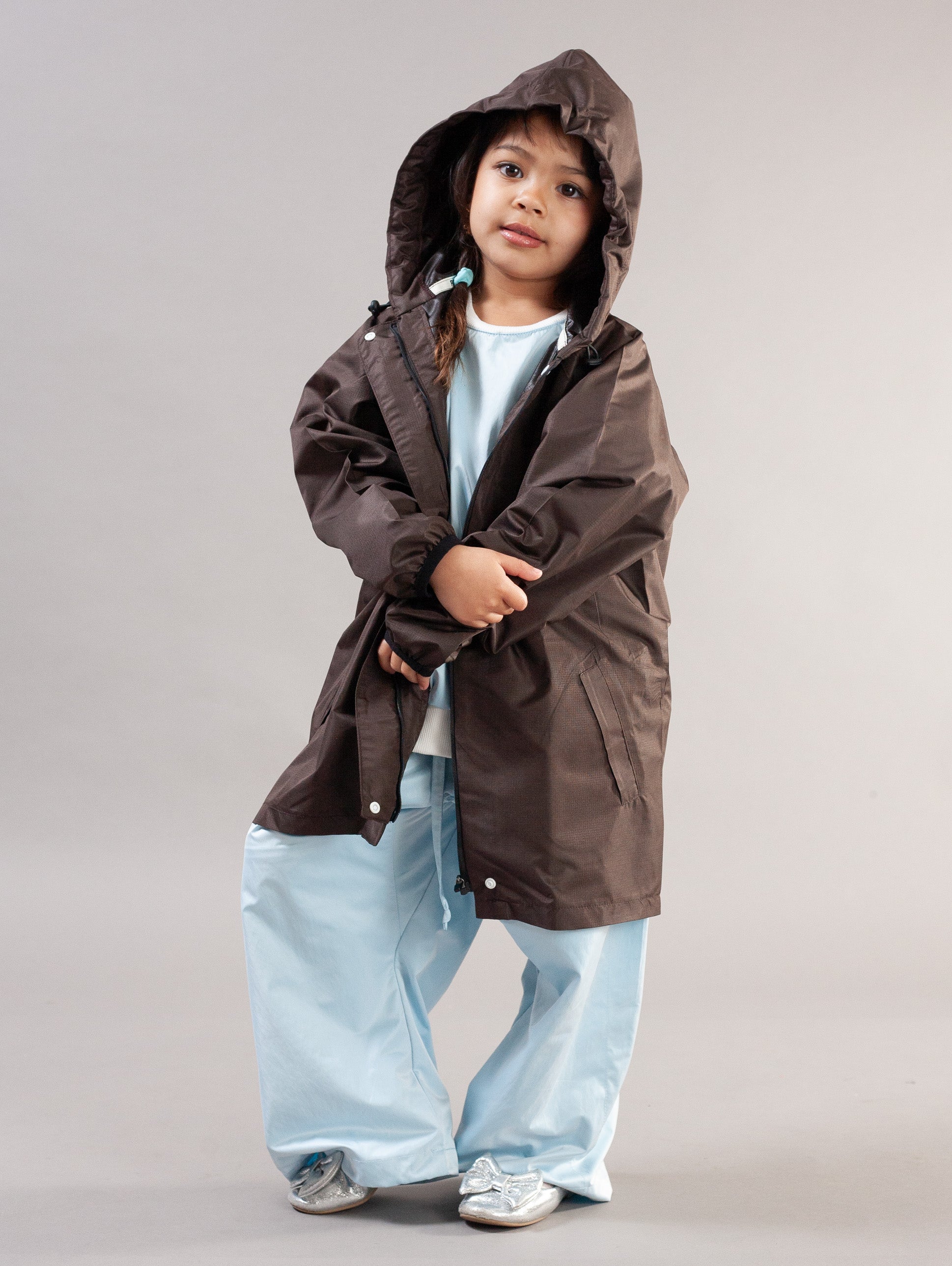 A young child wearing a dark brown hooded raincoat over a light blue shirt and wide-leg light blue pants, standing against a plain light gray studio background.