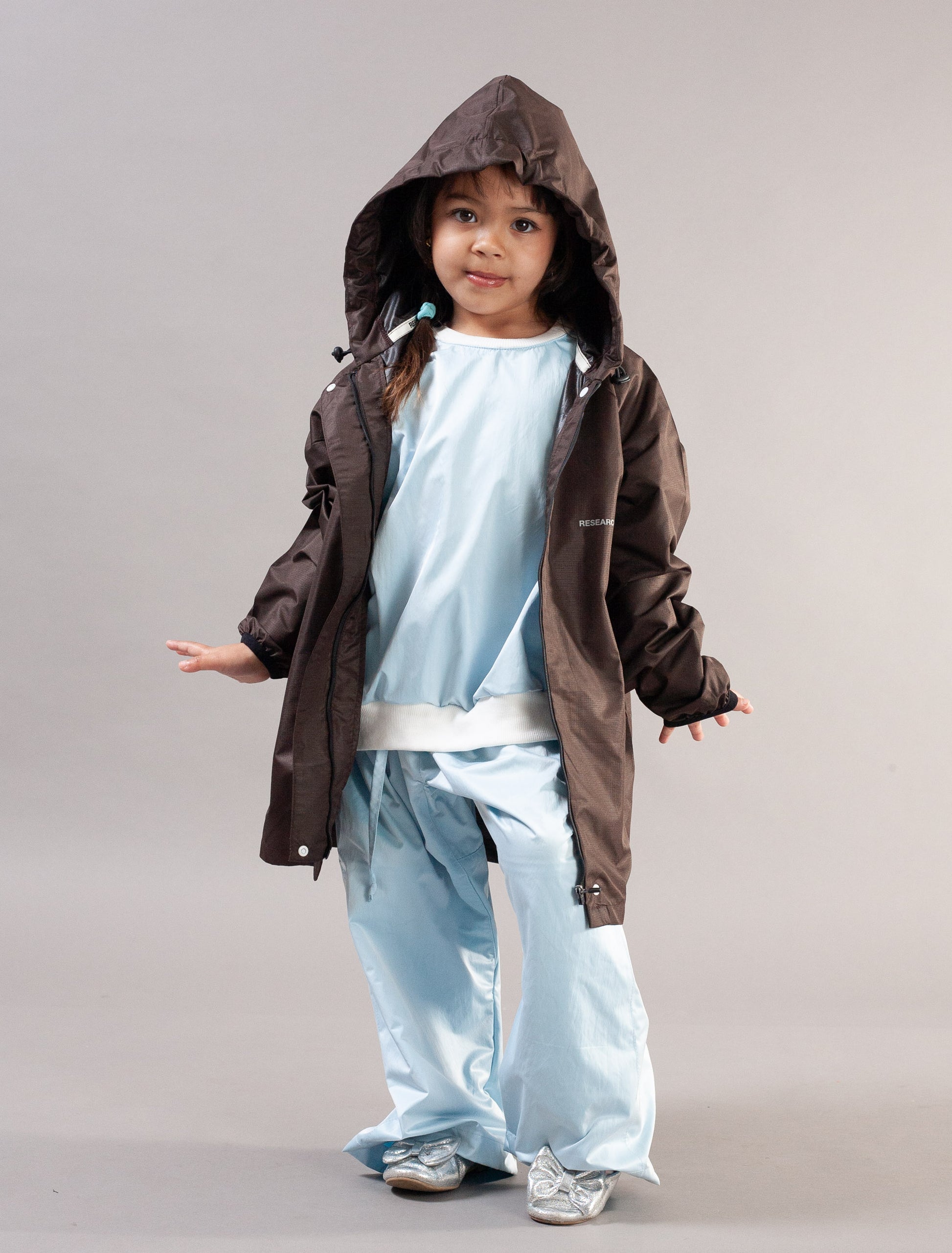 A young child modeling a dark brown hooded rain jacket over a light blue outfit, standing against a neutral gray studio background.