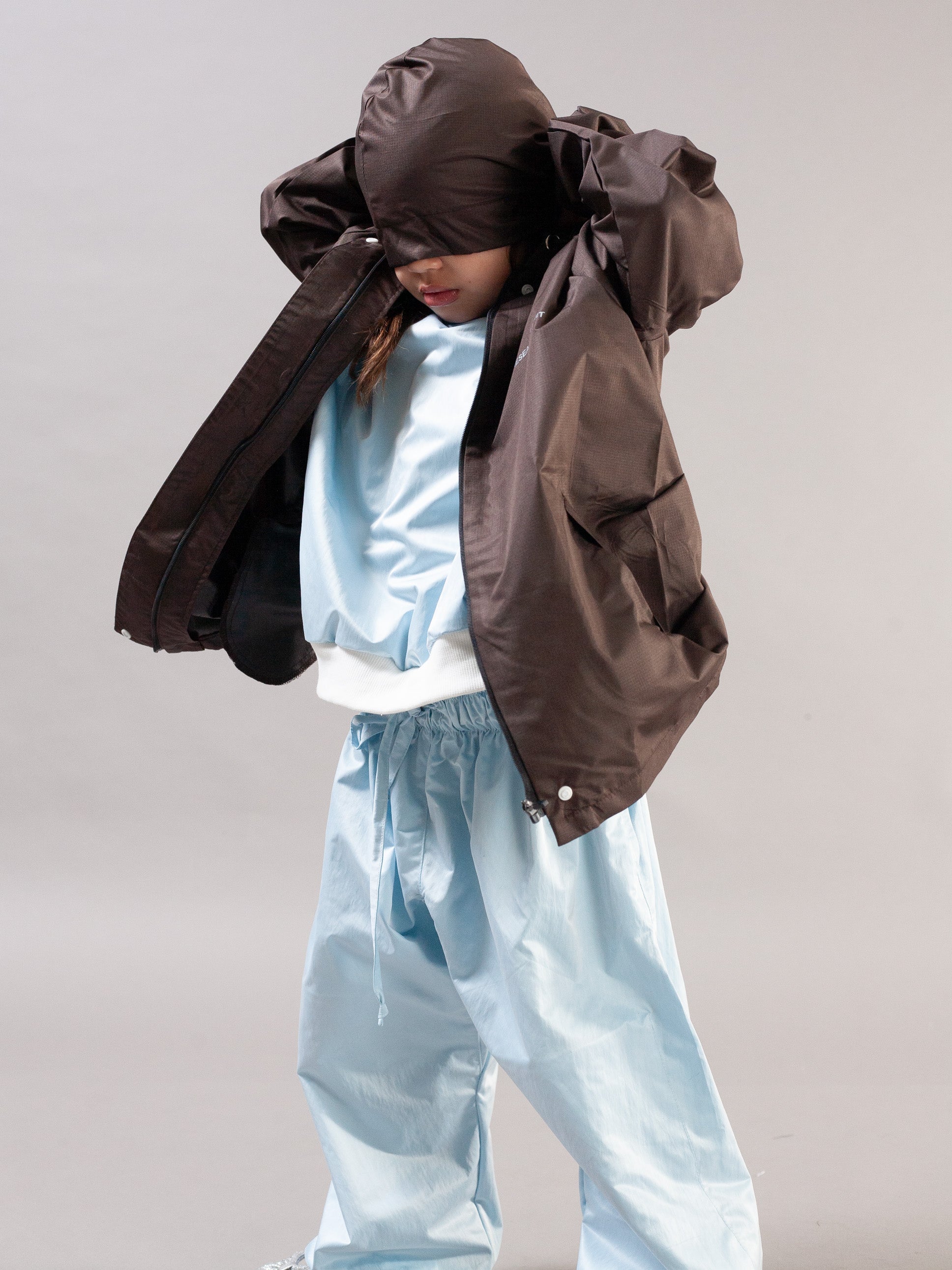 A child wearing a dark brown hooded jacket with the hood pulled over the eyes, layered over a light blue satin jacket and matching light blue baggy pants, against a plain gray studio background.