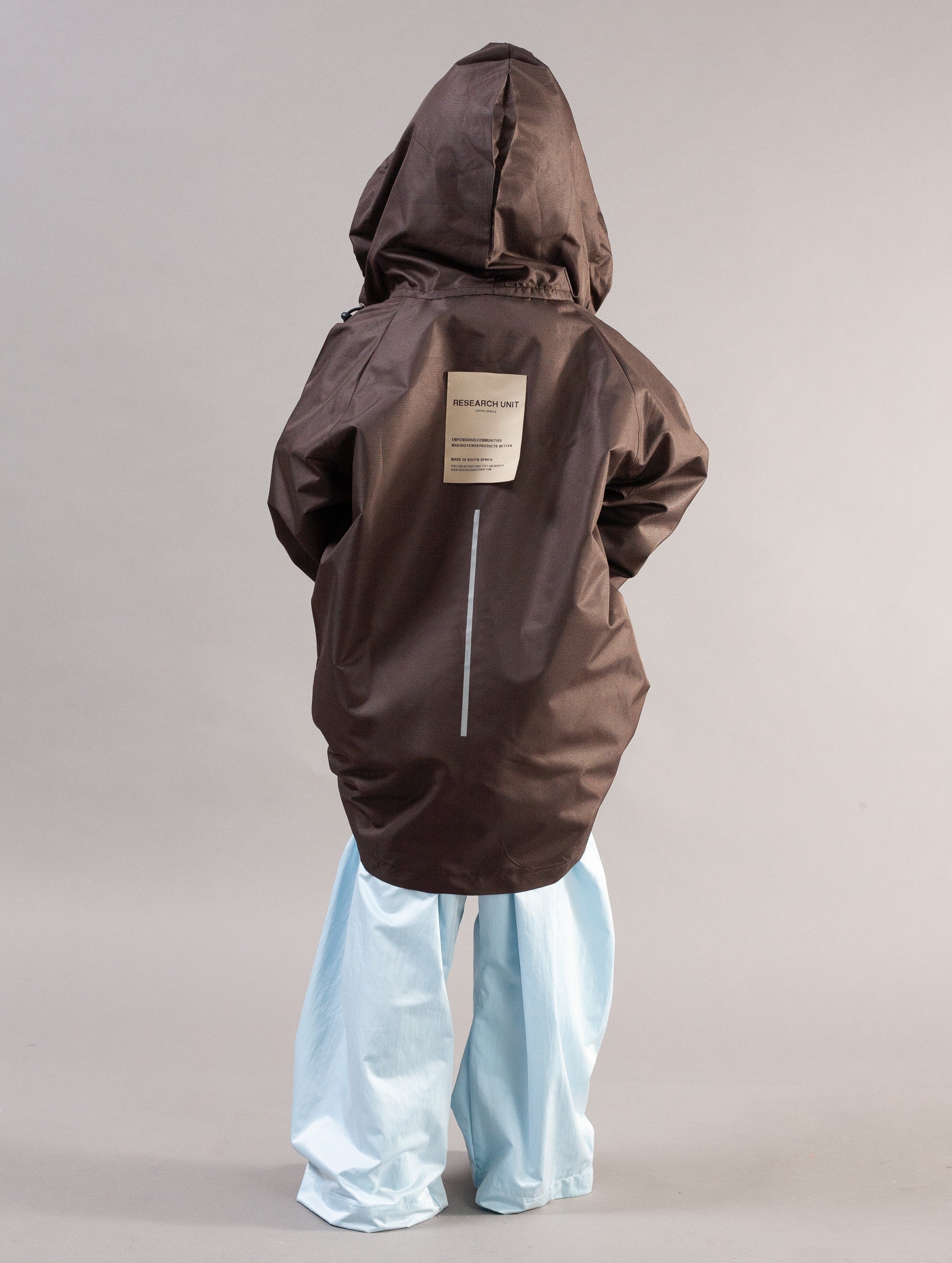 A child shown from the back wearing a dark brown hooded jacket with a light beige rectangular label on the back and light blue wide-leg pants, photographed against a plain light gray studio background.